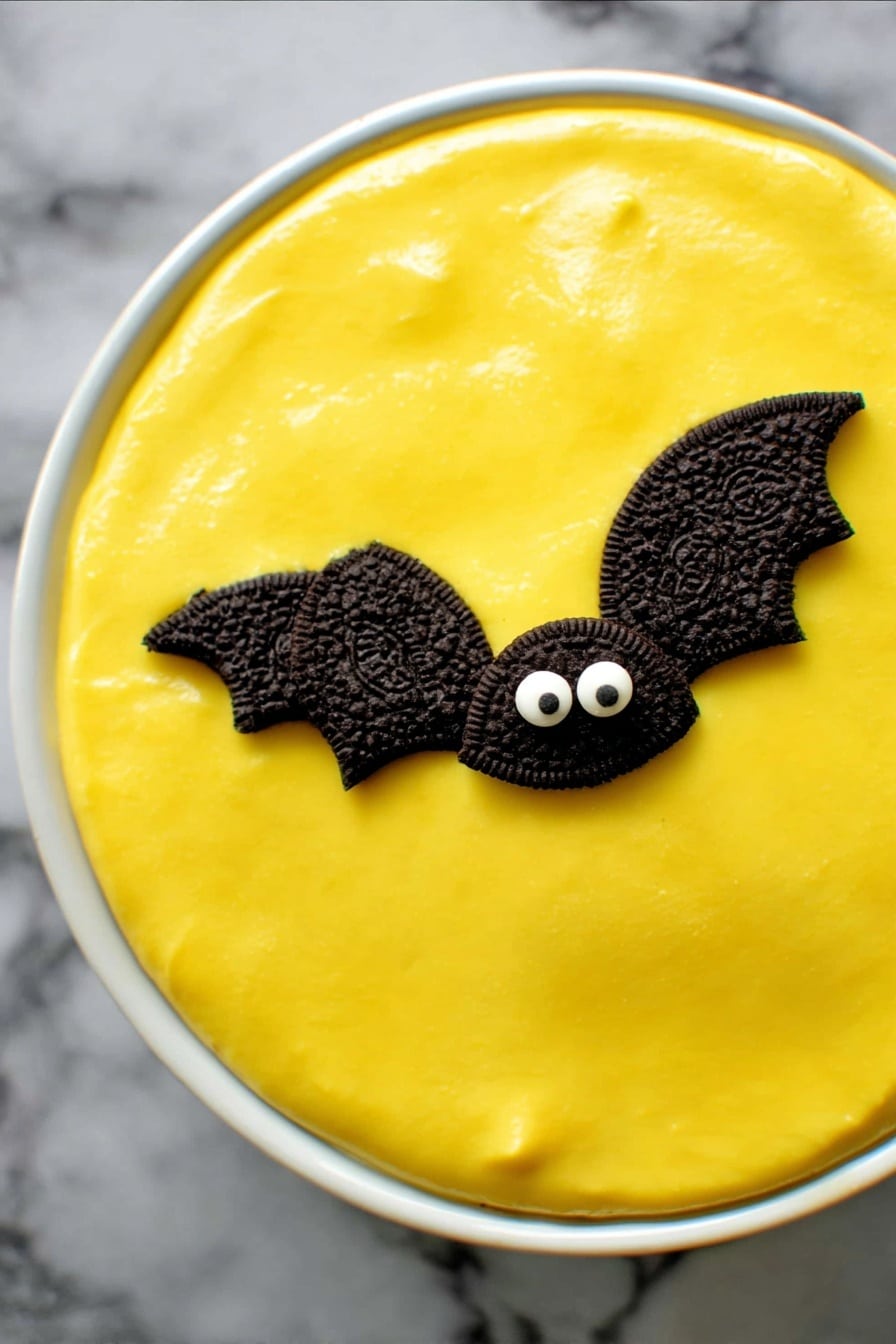 No Bake Boston Cream Halloween Pie, Halloween dessert, no bake Halloween pie, festive pumpkin pie, easy Halloween desserts - A round dessert in a white ceramic dish has a smooth, bright yellow top layer that looks creamy and shiny. On this yellow surface are two small decorations shaped like bats, made with halves of dark chocolate sandwich cookies for wings and two small white candy eyes on a chocolate base forming the face. The dish sits on a dark cloth background. photo taken with an iphone --ar 2:3 --v 7