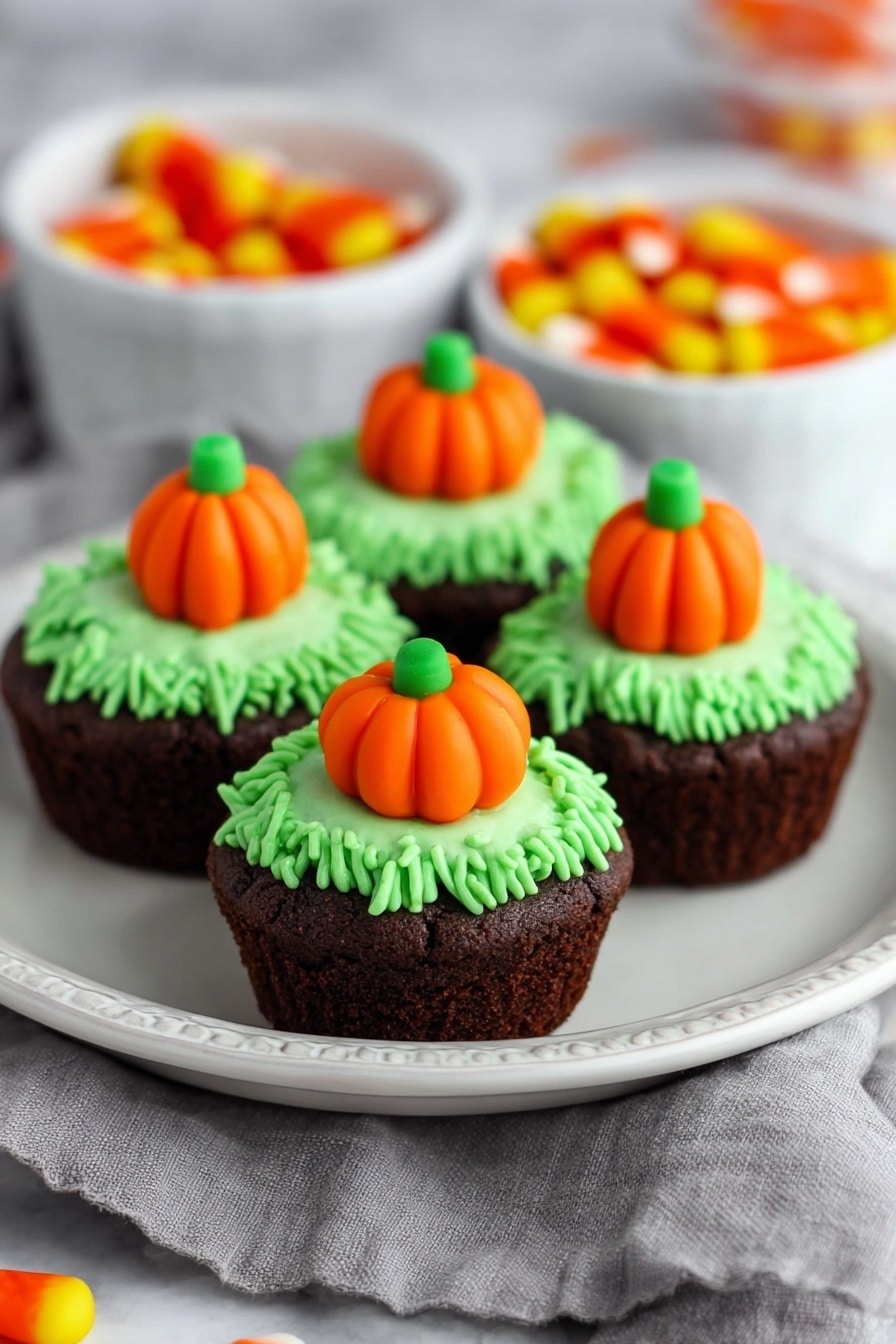 Pumpkin Patch Brownie Bites, fall dessert ideas, Halloween treats, festive brownie bites, pumpkin Halloween snacks - The image shows four small dark brown cupcakes placed closely on a white plate with a dotted edge. Each cupcake has a textured green frosting layer on top, piped to look like grass, and a small orange pumpkin-shaped candy with green detail in the center. The plate sits on a gray cloth on a white marbled surface. In the blurred background, two white bowls contain more orange pumpkin-shaped candies. The scene is bright with soft lighting highlighting the colors and textures. Photo taken with an iphone --ar 2:3 --v 7