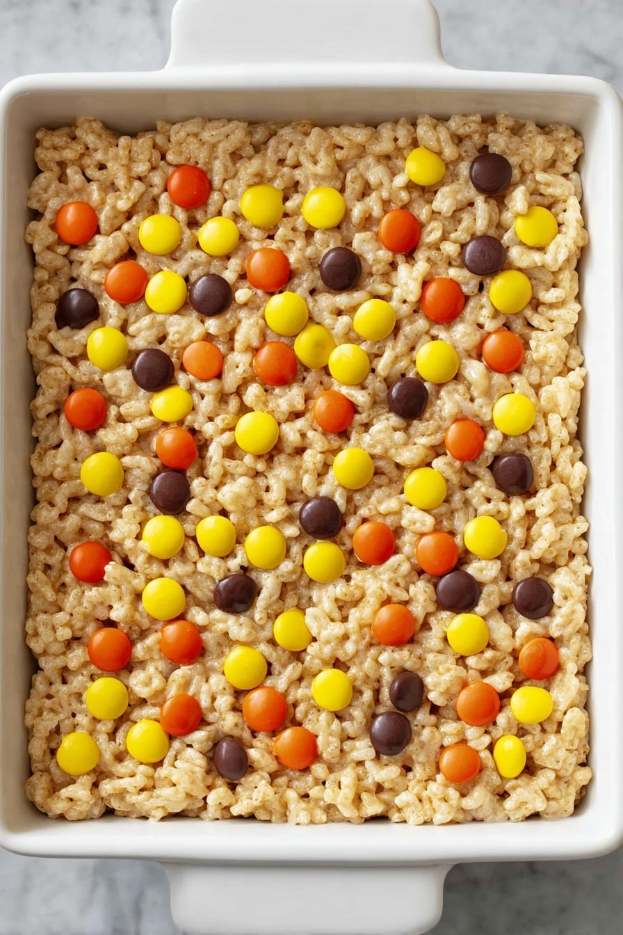 Chocolate Peanut Butter Rice Krispie Treats, peanut butter Rice Krispie bars, chocolate dessert recipes, easy Rice Krispie treats, no-bake chocolate peanut butter treats - A single thick layer of light golden crispy puffed rice cereal mixed with melted marshmallow is pressed into a white square dish, decorated on top with scattered small round candies in bright orange, yellow, and dark brown colors. The texture of the cereal layer looks soft and sticky but holds together, and the candies are evenly spread out on the surface against a white marbled background. Photo taken with an iphone --ar 2:3 --v 7