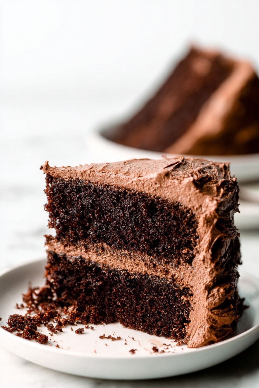 Gluten Free Vegan Chocolate Cake, gluten free vegan dessert, vegan chocolate cake, gluten free chocolate cake recipe, vegan cake without gluten - A close-up view of a two-layer chocolate cake slice on a white plate, placed on a white marbled surface. The cake layers are dark brown and moist with a rough texture. Between the layers is a medium brown chocolate frosting that looks creamy and smooth. The outside of the cake slice is covered in the same chocolate frosting, which is thick and slightly swirled, with rough edges. Some frosting has spilled onto the plate. In the background, there is a blurred image of another chocolate cake slice on a white plate. Photo taken with an iphone --ar 2:3 --v 7