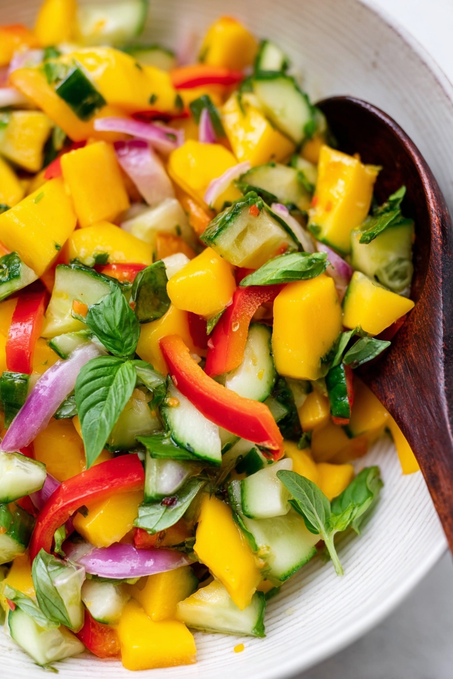 Mango Salad with Lemon-Honey Dressing, mango salad recipe, refreshing fruit salad, easy summer salad, healthy mango salad - A close-up of a colorful salad in a white bowl with a white marbled background, showing bright yellow mango cubes, green cucumber pieces, red bell pepper chunks, thin slices of light purple onion, and fresh green basil leaves mixed together. A dark wooden spoon is resting inside the bowl, partly covered by the salad. The colors are vibrant and fresh, with the textures of the juicy mango, crunchy cucumber, and leafy greens clearly visible. Photo taken with an iphone --ar 2:3 --v 7