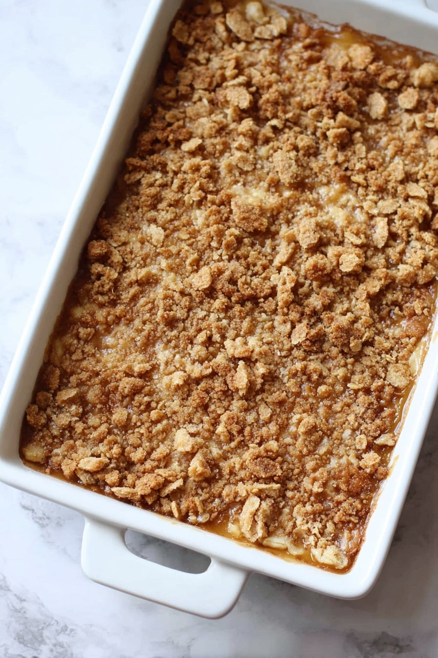 Pumpkin Dump Cake, easy pumpkin dessert, fall dessert recipes, quick pumpkin cake, pumpkin comfort food - The image shows a baked dessert in a white rectangular dish with two handles. The dessert has one main layer with a golden brown crumbly topping made of crushed pieces with a rough, uneven texture. The color is mixed shades of light to medium brown, giving a crunchy and toasted look. The dish is placed on a white marbled surface. Photo taken with an iphone --ar 2:3 --v 7