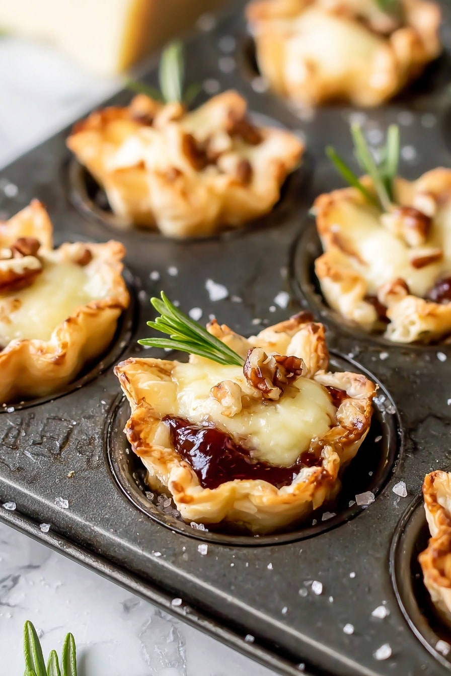 Cranberry Brie Bites with Puff Pastry, cranberry brie appetizer, holiday puff pastry bites, easy crowd-pleasing appetizers, festive party snacks - The image shows mini tartlets baked in a dark metal muffin tray, each tartlet having three main layers. The bottom layer is a golden-brown, flaky pastry shaped into small cups with slightly crispy edges. The middle layer is a glossy, dark red jam or fruit preserve that peeks out from beneath the top. The top layer is a melted, creamy pale yellow cheese, topped with small brown nut pieces and a fresh green rosemary sprig for garnish. The background is a white marbled texture, and some salt granules are scattered around the tray. photo taken with an iphone --ar 2:3 --v 7