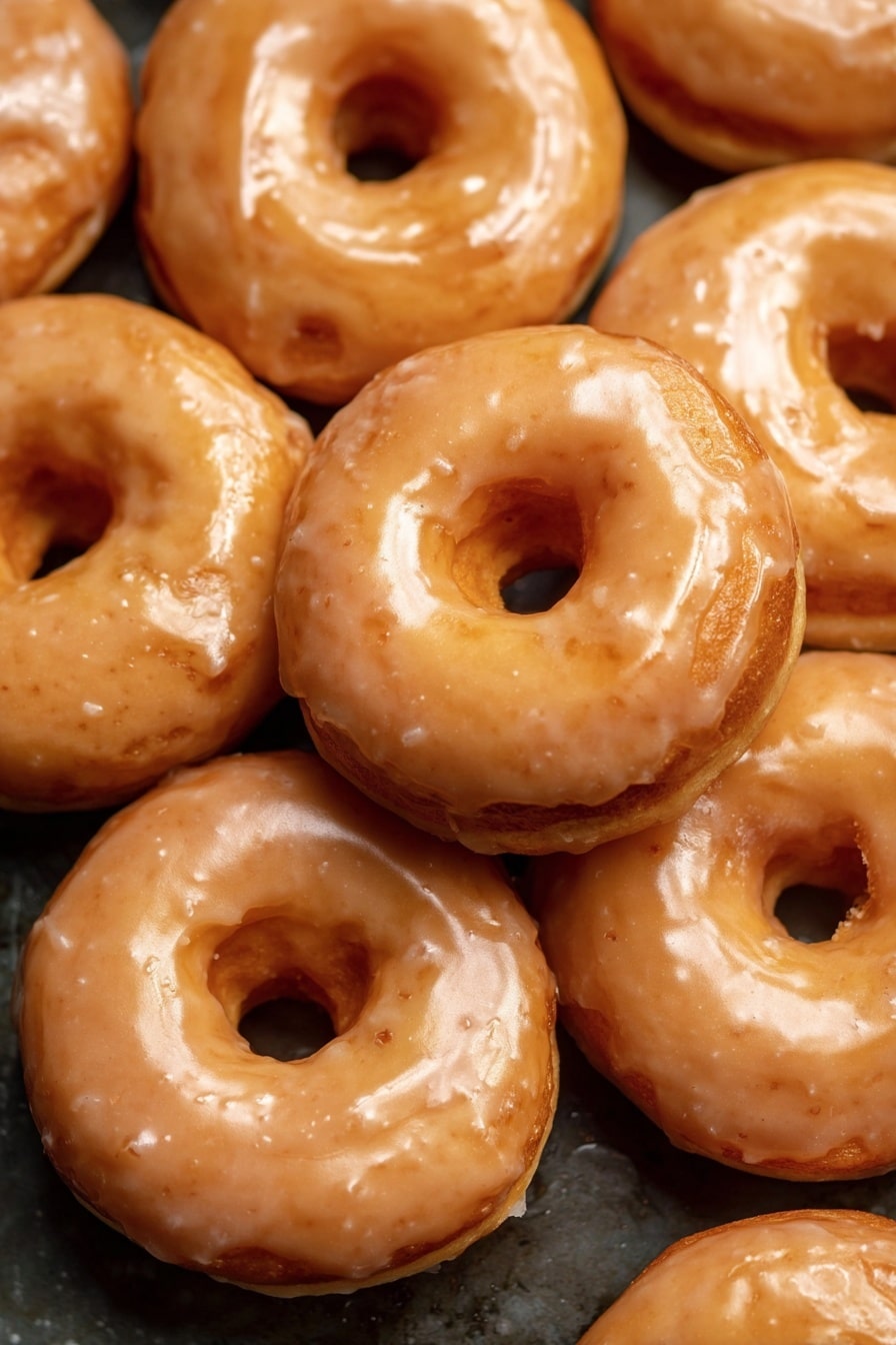 Vegan Pumpkin Donuts with Cinnamon Maple Glaze, vegan pumpkin donut recipe, fall vegan donuts, pumpkin spice donuts, plant-based pumpkin treats - The image shows a close-up of many round donuts stacked together. Each donut has two visible layers: a base layer that is a golden brown color with a soft texture and a shiny glaze layer on top that is light tan and slightly translucent. The glaze evenly covers the top and sides of each donut, giving them a glossy look. The donuts have a round hole in the middle, and their surface shows slight bumps and smooth spots. The background is a dark surface contrasting with the shiny donuts. photo taken with an iphone --ar 2:3 --v 7