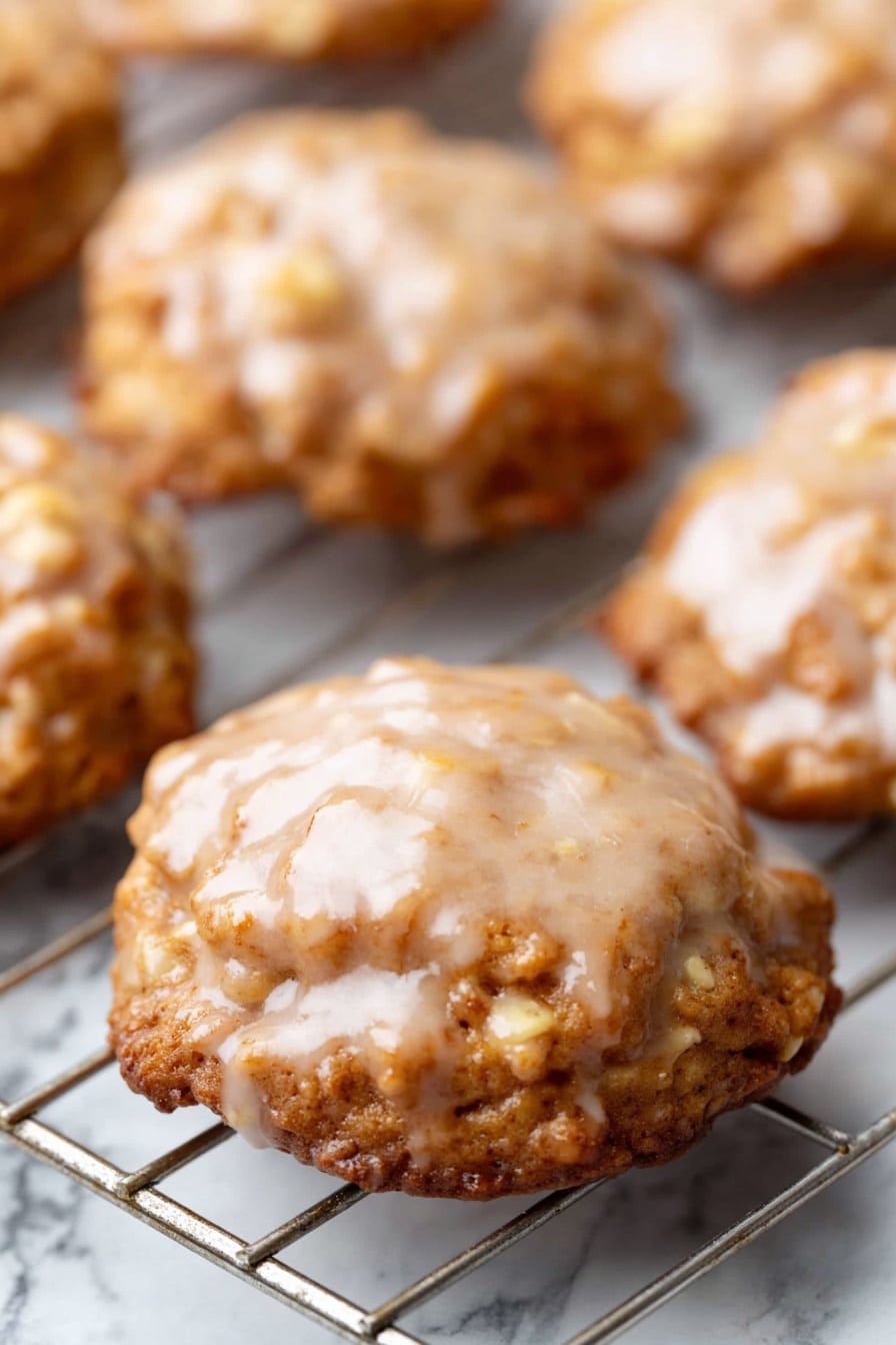Crispy Apple Fritters with Sweet Vanilla Glaze, apple fritter recipe, easy apple fritters, homemade apple fritters, crunchy apple treats - The image shows several round cookies on a cooling rack. Each cookie has a shiny, light brown glaze on top with a slightly uneven texture. The cookies have a golden brown color, with a rough surface and visible small pieces inside, likely nuts or oats. The cooling rack underneath is metal and placed over a white marbled surface. The cookies look soft and have a slightly wavy edge. Photo taken with an iphone --ar 2:3 --v 7