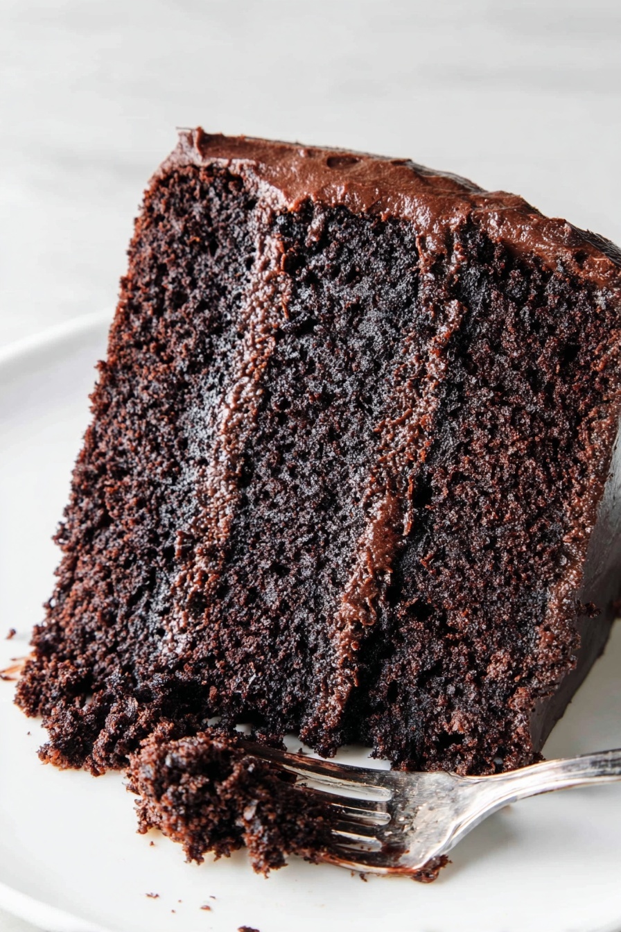 The Most Amazing Chocolate Cake, moist chocolate cake recipe, homemade chocolate cake, easy chocolate cake, rich chocolate dessert - The image shows a slice of chocolate cake on a white plate placed on a white marbled surface. The cake has two thick layers of moist, dark brown chocolate sponge with a rich, glossy chocolate frosting layer in the middle. The outside of the cake is covered with a smooth, shiny chocolate frosting. A silver fork with some chocolate frosting rests on the plate next to the slice. Photo taken with an iphone --ar 2:3 --v 7
