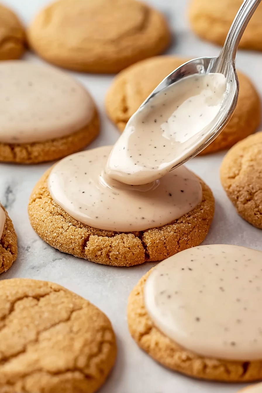Brown Sugar Pop Tart Cookies, homemade pop tart cookies, easy dessert recipes, cinnamon sugar cookies, sweet snack ideas - Round light brown cookies with a soft, cracked surface are arranged on a white marbled background. A silver spoon is shown spreading a smooth, pale beige icing with small dark specks on the top surface of one cookie near the center. Several other cookies already have the same thin layer of icing evenly covering their tops while some remain plain. The cookies are placed closely together in rows and the icing appears glossy and creamy. Photo taken with an iphone --ar 2:3 --v 7