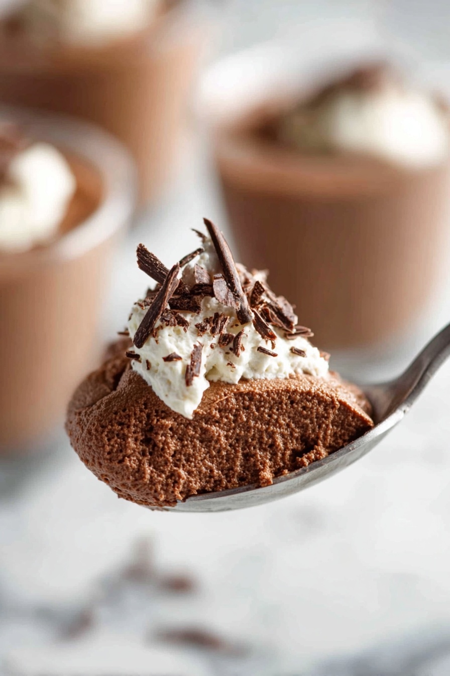 Decadent Chocolate Mousse, chocolate mousse recipe, rich chocolate dessert, airy chocolate mousse, homemade chocolate mousse - The image shows a clear glass filled with a rich, smooth dark brown chocolate pudding. On top, there is a dollop of white whipped cream with delicate dark chocolate shavings scattered over it. The glass is placed on a white marbled surface, and in the background, there are blurred similar glasses filled with the same dessert. Photo taken with an iphone --ar 2:3 --v 7
