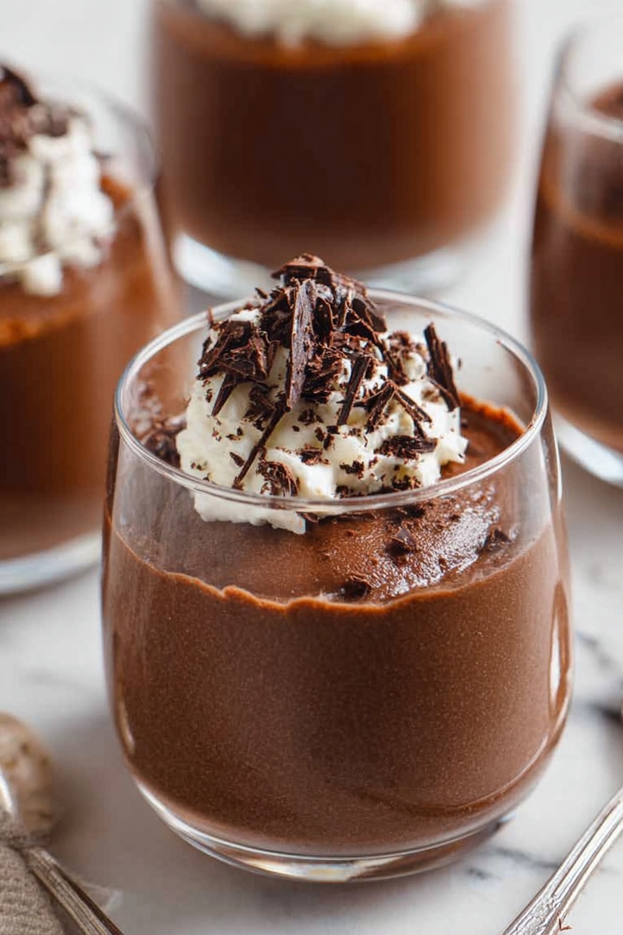 Decadent Chocolate Mousse, chocolate mousse recipe, rich chocolate dessert, airy chocolate mousse, homemade chocolate mousse - A close-up of a spoon holding a scoop of light and airy chocolate mousse with a rough texture, topped with a small dollop of white whipped cream and thin, dark chocolate shavings scattered on top, with two blurry white bowls filled with the same mousse and cream in the background, all set on a white marbled surface. photo taken with an iphone --ar 2:3 --v 7