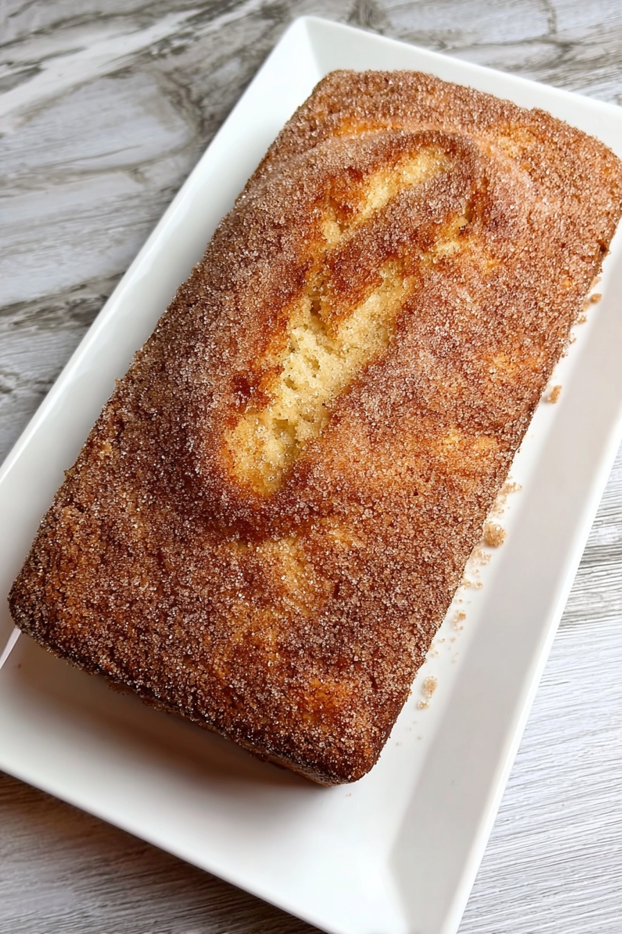 Cinnamon Donut Bread, easy cinnamon bread, soft cinnamon loaf, cinnamon sugar bread, comforting baked bread - A rectangular cake with a golden-brown top layer covered evenly with a fine, grainy light brown sugar coating sits on a white rectangular plate. The cake's top surface shows a gently cracked texture with soft ridges and swirls, revealing a tender and moist interior below the sugar crust. The plate rests on a white marbled texture surface. photo taken with an iphone --ar 2:3 --v 7