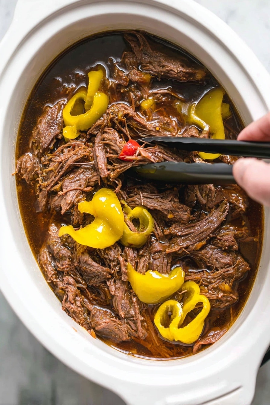 Italian Slow Cooker Beef Sandwiches with Pepperoncini, beef sandwich recipes, slow cooker beef sandwiches, pepperoncini beef sandwich ideas, easy slow cooker sandwiches - A close-up shot of shredded brown meat soaked in dark brown juices inside a white oval slow cooker. The meat is mixed with bright yellow sliced peppers and whole pepper shapes on top. Woman's hand is visible holding black tongs, lifting some meat from the cooker. The background surface is white marble. photo taken with an iphone --ar 2:3 --v 7