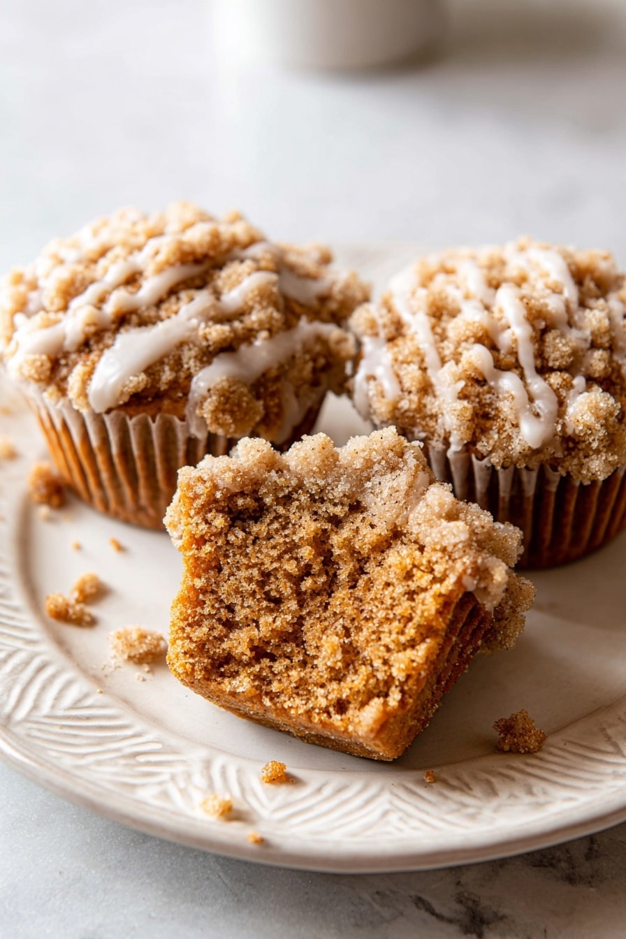 Pumpkin Crumb Cake Muffins, pumpkin muffins with crumb topping, fall baking recipes, easy pumpkin muffin recipe, cozy autumn desserts - The image shows seven golden brown muffins arranged closely on a white plate. Each muffin has a crumbly, rough-textured top layer that is light brown and covered with a light cream-colored glaze that drips down the sides in thin streaks. The muffins are placed on a smooth white marbled surface underneath the plate. Some crumbs are scattered around the plate and surface. The lighting highlights the texture and glaze shine on the muffins. Photo taken with an iphone --ar 2:3 --v 7