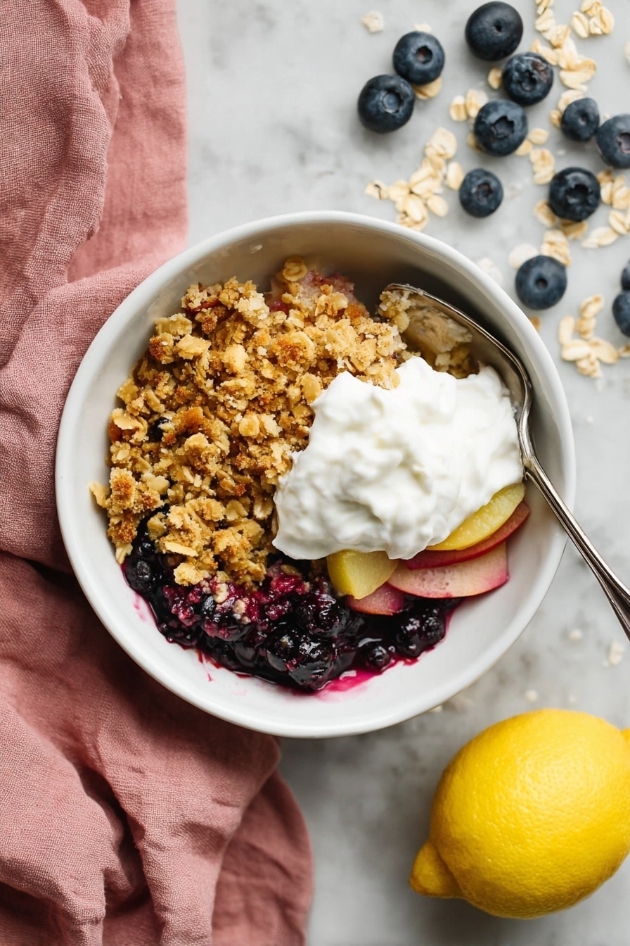Apple and Blueberry Crumble, Apple and Blueberry Crumble Recipe, easy fruit crumble dessert, homemade fruit crumble, cozy fall dessert - A white bowl sits on a white marbled surface, filled with three main layers. The bottom layer contains dark blue and purple berries mixed with thin slices of light pink fruit. Above that is a thick, crumbly layer of golden brown oat crumble with hints of toasted oats on top. The final layer is a generous dollop of white whipped cream placed in the center of the crumble. A silver spoon is partly inside the bowl on the right side. Next to the bowl is a yellow lemon and scattered blueberries and oats, with a soft pink cloth to the left. photo taken with an iphone --ar 2:3 --v 7