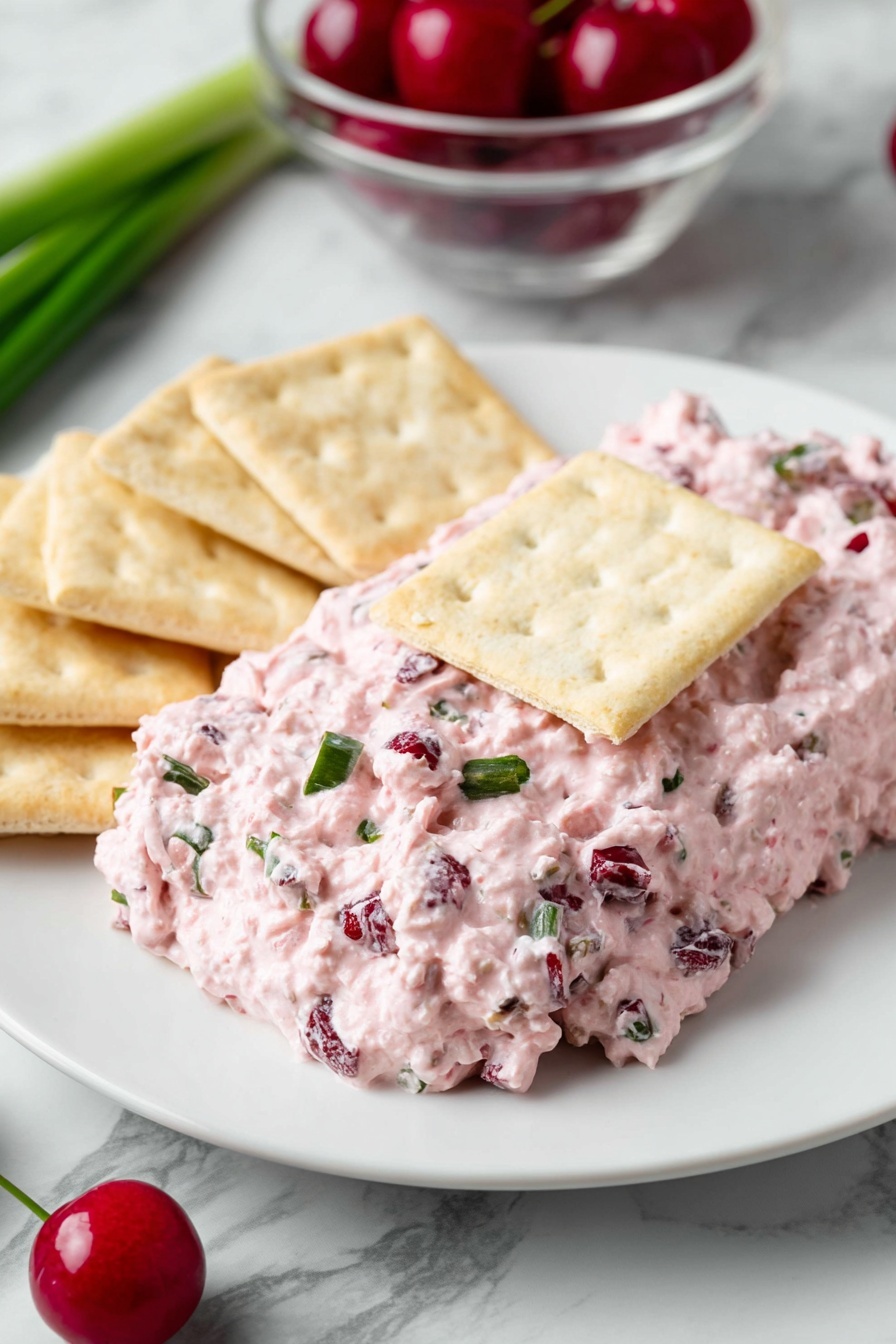 Cranberry Jalapeno Dip, holiday dip with cranberries and jalapenos, spicy and sweet dip, easy vegan dip recipes, festive party dip - On a white plate, there is a thick layer of pink creamy spread with small chunks of darker red and green mixed in. The spread is topped with a rectangular light beige cracker in the center. Around the spread, there are more light beige crackers placed on the white plate. The background shows a green onion on a white marbled surface and a clear bowl with red cherries. A green chili pepper is partially visible near the plate. photo taken with an iphone --ar 2:3 --v 7