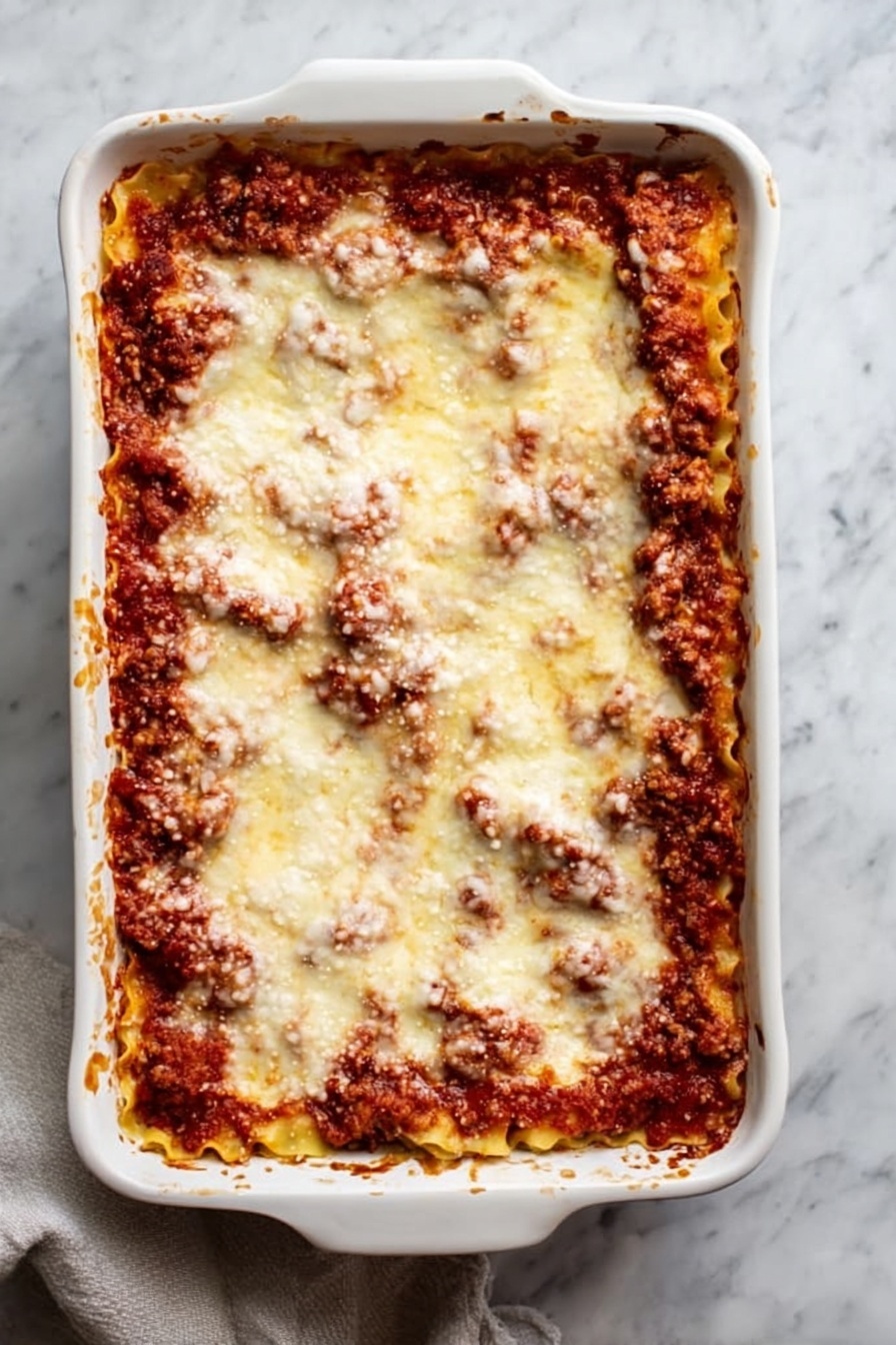 Vegan Lasagna Rolls, vegan lasagna rolls recipe, plant-based lasagna rolls, vegan lasagna roll-ups, vegan pasta casserole - A white rectangular ceramic baking dish filled with a baked lasagna, showing at least three layers: a bottom layer of pasta sheets, a middle layer of rich red tomato sauce mixed with browned ground meat, and a top layer covered evenly with melted, slightly browned cheese that looks soft and smooth. The edges of the dish have some browned sauce splashes. The dish sits on a surface with a white marbled texture, and a light-colored cloth is partially visible under the dish on the lower left side. Photo taken with an iphone --ar 2:3 --v 7