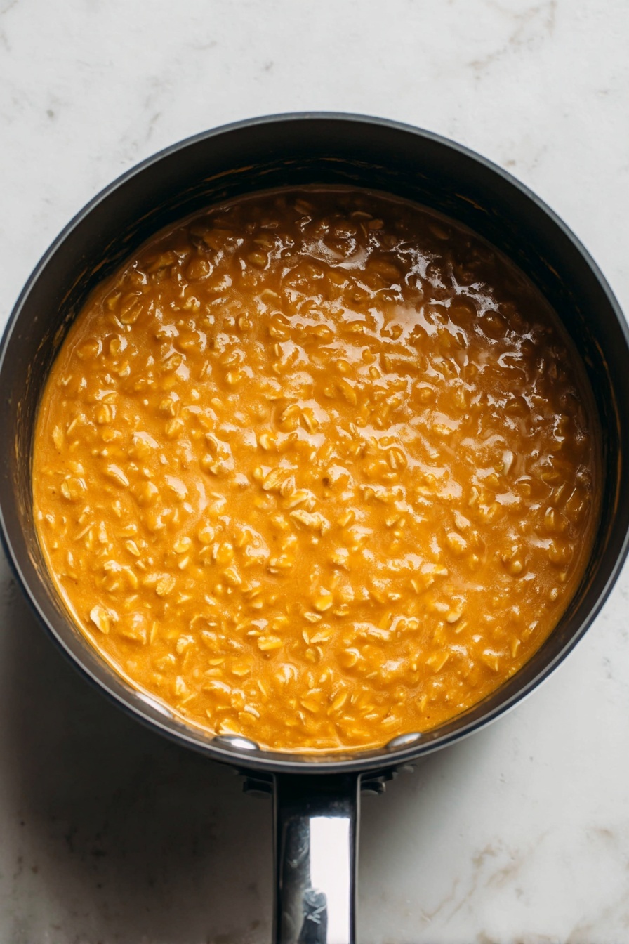 Pumpkin Oatmeal, Pumpkin Oatmeal Recipe, Fall Breakfast Ideas, Cozy Morning Breakfast, Healthy Pumpkin Breakfast - This image shows a close-up top view of a round black saucepan filled with a thick, creamy orange mixture with visible small oats throughout. The oats give the surface a bumpy texture, and the mixture looks smooth and warm. The saucepan is placed on a white marbled surface, and the shiny black handle is visible at the bottom right of the image. The lighting highlights the glossy shine of the mixture, making it look fresh and appetizing. photo taken with an iphone --ar 2:3 --v 7