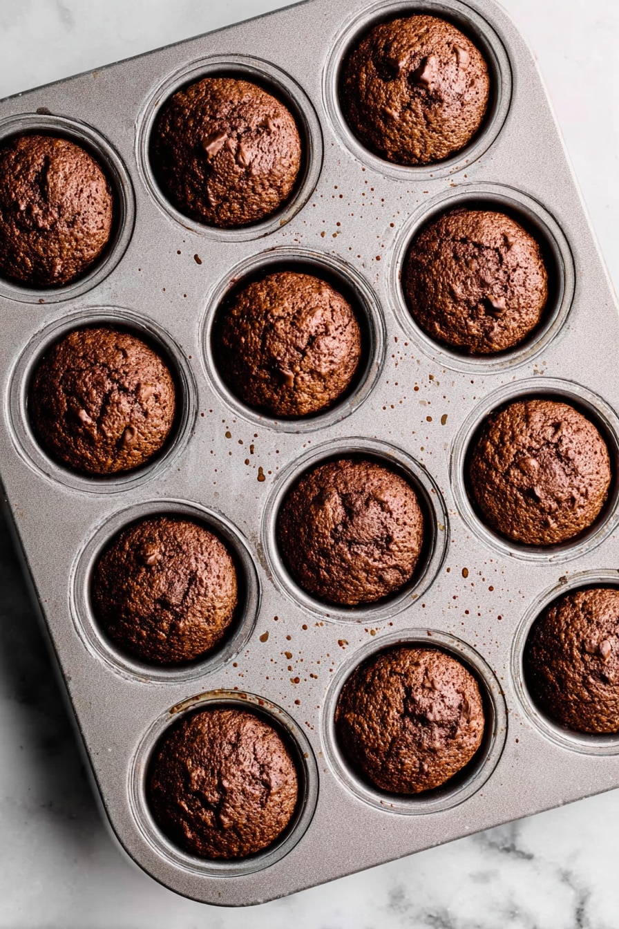 Sticky Toffee Pudding with Warm Toffee Sauce, moist toffee pudding recipe, easy sticky toffee dessert, warm toffee sauce recipe, comforting caramel pudding - A metal muffin tray with 12 evenly spaced round muffins baked inside, each muffin a deep brown color with a rough, slightly cracked top texture showing some small chunks within. The muffins fit neatly in each circular slot of the tray, which shows small spots of light brown residue from baking. The tray is placed on a white marbled surface with soft natural lighting highlighting the muffins' golden and dark brown hues. photo taken with an iphone --ar 2:3 --v 7