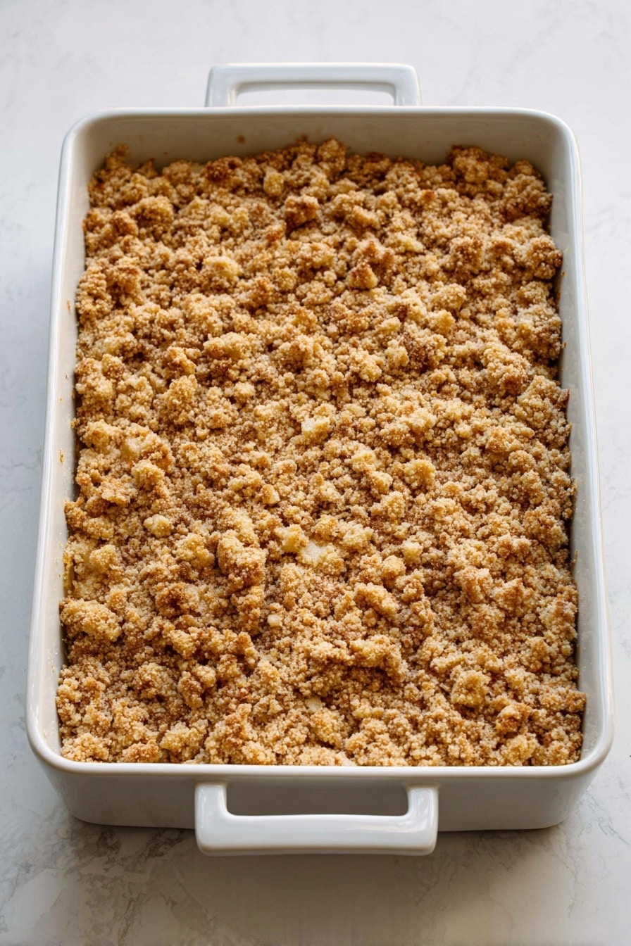 Caramel Apple Cheesecake Bars, caramel apple dessert, fall dessert recipes, apple cheesecake bars, easy cheesecake bar recipe - A white rectangular baking dish with two handles is filled with a golden-brown crumbly topping that looks crunchy and uneven in texture, covering the entire surface. The topping has small clusters and a slightly rough look with hints of light and darker toasted shades. The dish is placed on a white marbled surface. photo taken with an iphone --ar 2:3 --v 7