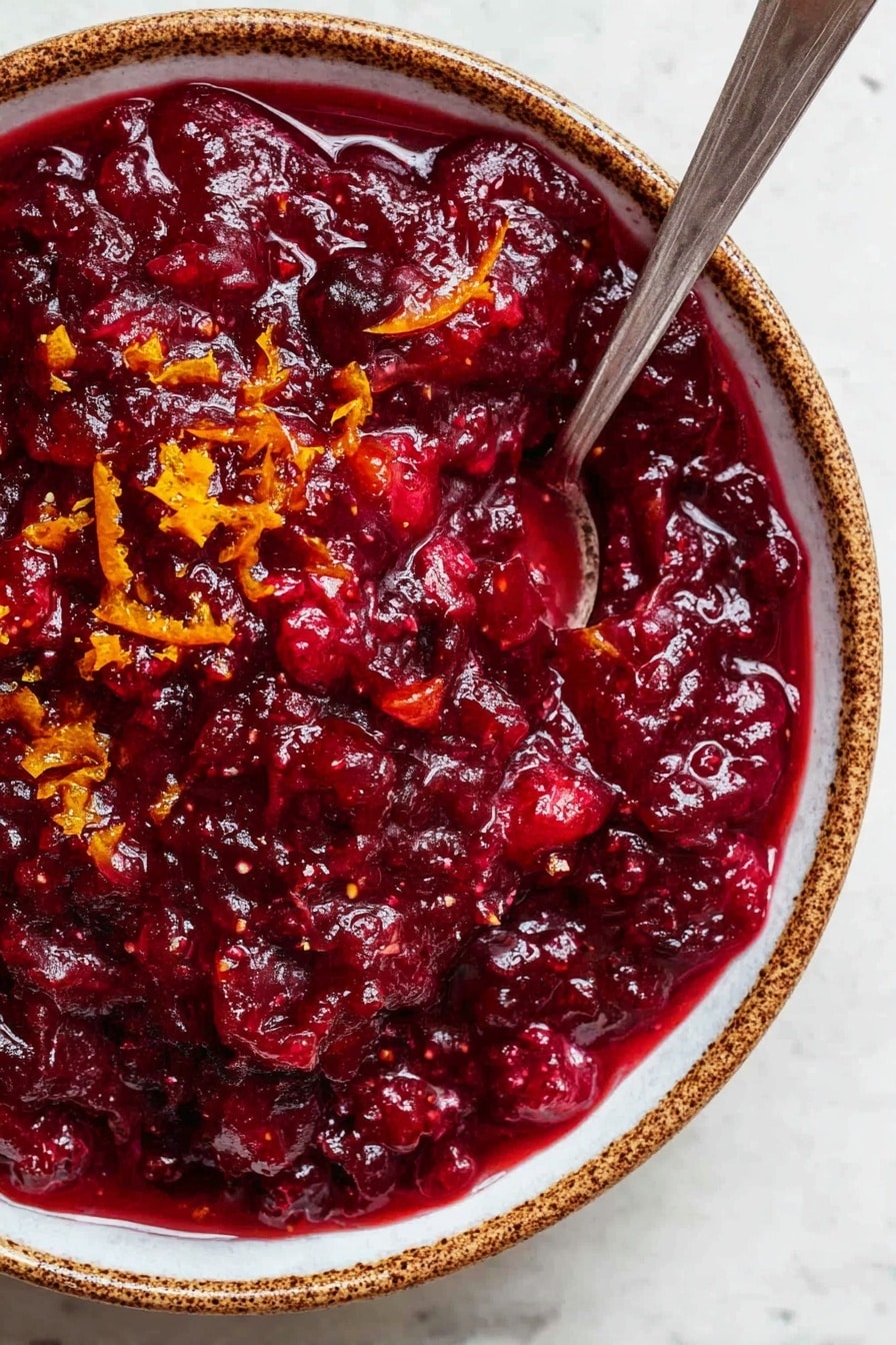 Easy Cranberry Sauce with Orange Juice, cranberry sauce recipes, homemade cranberry sauce, orange-infused cranberry sauce, holiday cranberry sauce - A close-up view of a thick, deep red cranberry sauce with a chunky texture, showing visible pieces of fruit mixed in. Small bits of bright orange zest are scattered on top, adding color contrast. The sauce fills a white bowl with brown speckled edges, and a silver spoon is placed inside the bowl, partly submerged in the sauce. The bowl sits on a white marbled surface. photo taken with an iphone --ar 2:3 --v 7