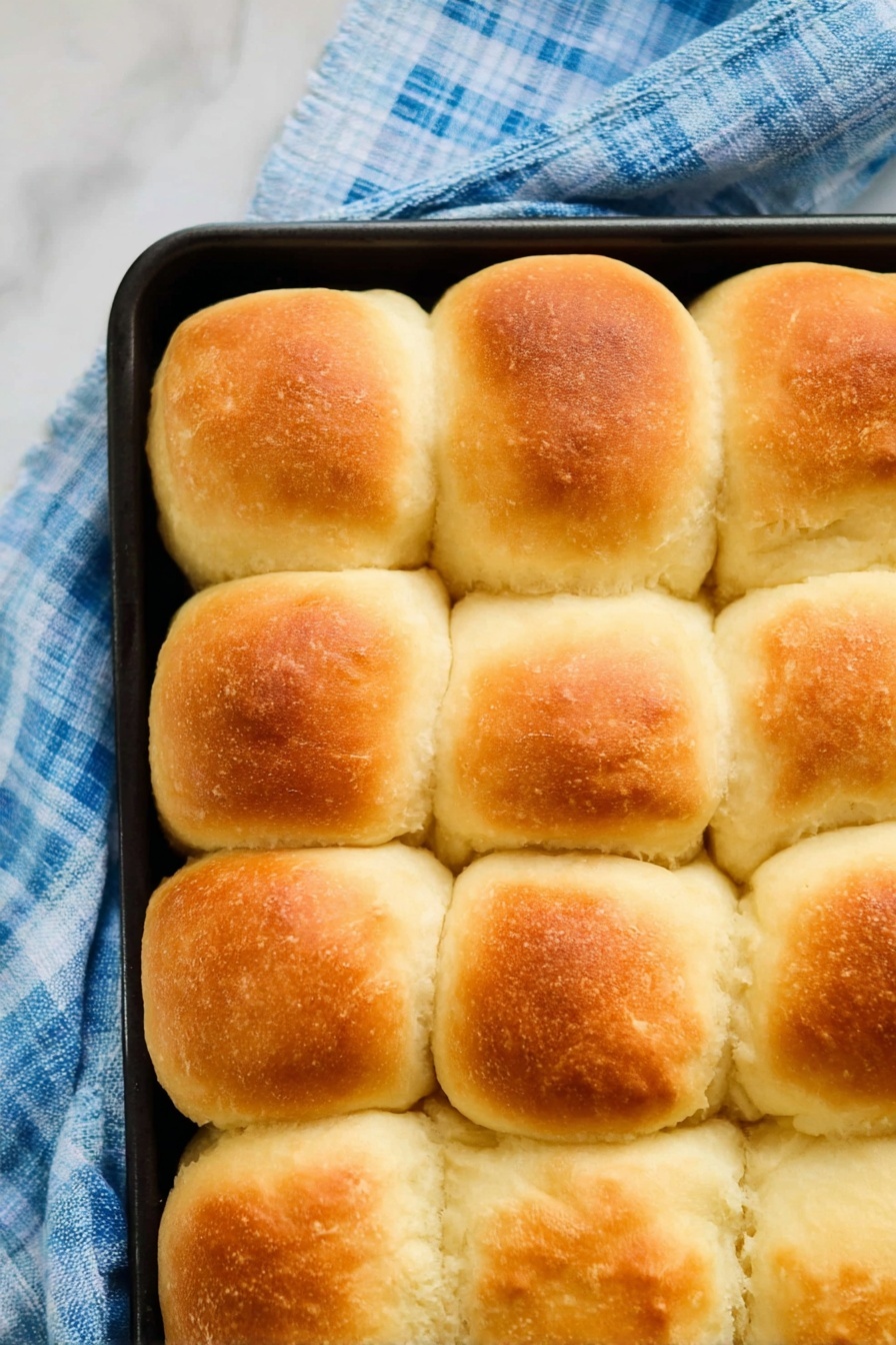 Vegan Dinner Rolls, vegan bread rolls, fluffy plant-based dinner rolls, homemade vegan buns, easy vegan bread - The image shows a close-up top view of sixteen golden-brown, fluffy bread rolls arranged in a 4 by 4 grid inside a black baking tray. Each roll has a slightly rounded top with a soft, smooth texture and a light crust that is golden on the upper part, fading to creamy white around the sides. The baking tray is placed on a blue and white checkered cloth on a white marbled surface. The rolls touch each other, showing their soft and puffy sides. photo taken with an iphone --ar 2:3 --v 7