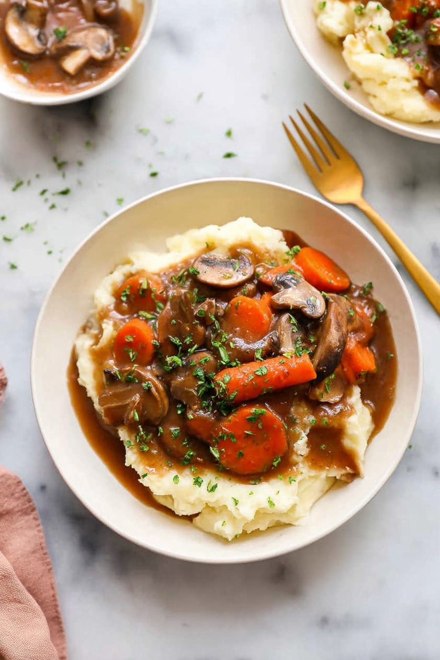 Mushroom Bourguignon, vegetarian mushroom stew, plant-based Bourguignon, French mushroom stew, hearty mushroom dinner - A white bowl holds a dish with two main layers: the bottom layer is creamy mashed potatoes, soft and pale in color, covering the base of the bowl. On top is a rich brown gravy mixed with sliced mushrooms and large carrot chunks, with chopped green herbs sprinkled all over for color. A golden spoon and fork sit on the white marbled surface beside the bowl. In the background, another white bowl with the same dish is barely visible. The lighting is bright and natural, highlighting the textures and colors of the food photo taken with an iphone --ar 2:3 --v 7