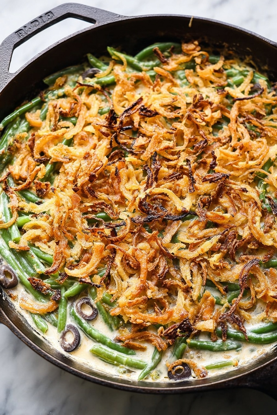 Vegan Green Bean Casserole, plant-based green bean casserole, dairy-free green bean side dish, vegan holiday side recipe, crispy fried onion green beans - This image shows a close-up of a baked green bean casserole in a cast iron pan placed on a white marbled surface. The bottom layer consists of bright green beans mixed with sliced black olives visible within a creamy, white sauce. On top of this, there is a thick, even layer of crispy, golden-brown fried onion strips spread across the entire pan, giving the dish texture and a crunchy appearance. Some onion strips have darker, almost burnt edges which add color contrast. The round pan's black handle is partly visible, framing the dish. Photo taken with an iphone --ar 2:3 --v 7