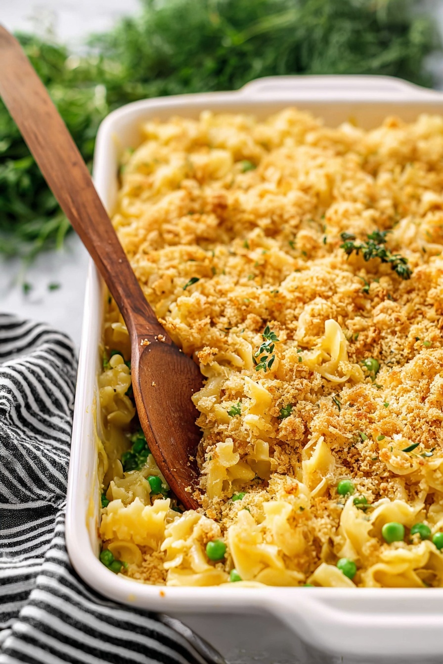 Creamy Chicken Casserole, Chicken casserole recipe, cheesy chicken bake, comfort food dinner, easy chicken casserole - The image shows a white rectangular baking dish filled with a pasta casserole. The dish has three main layers: a bottom layer of yellow pasta, a middle layer with green peas mixed throughout, and a top layer covered with a golden brown crumbly topping that looks crispy. A wooden spoon stands inside the dish, scooping some pasta and crumbs. The background features a white marbled surface and some green herbs with a black and white striped cloth nearby. photo taken with an iphone --ar 2:3 --v 7