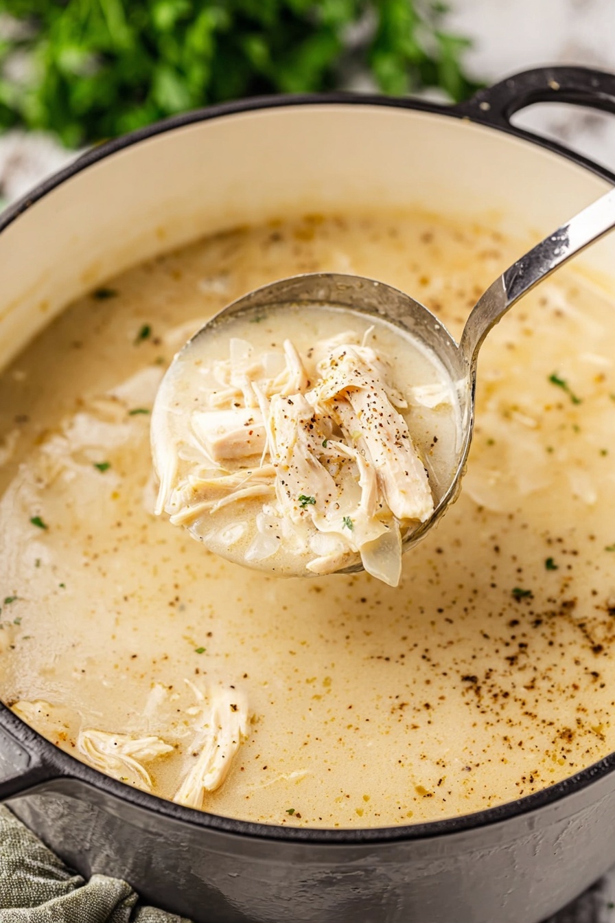 Southern Style Chicken and Dumplings, chicken and dumplings recipe, comforting Southern chicken dish, homemade chicken and dumplings, easy chicken soup with dumplings - A close-up view of a creamy light beige soup with shredded light tan chicken pieces mixed in. The soup has a smooth, thick texture with visible specks of black pepper spread on the surface. A shiny metal ladle lifts a portion of the soup showing the creamy broth and chicken clearly. The soup pot is white inside with a black exterior handle visible, resting on a white marbled surface with green blurred herbs in the background. Photo taken with an iphone --ar 2:3 --v 7