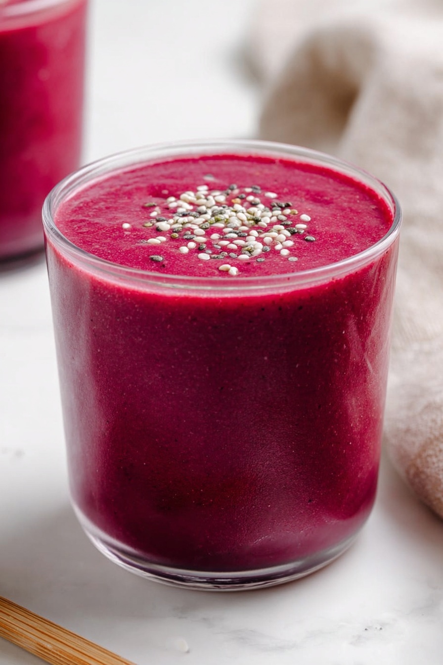 Banana Beet Smoothie, healthy beet smoothie, nutritious fruit smoothie, vibrant green smoothie, easy smoothie recipes - A clear glass is filled with a thick, vibrant dark pink smoothie, almost reaching the top with a smooth and creamy texture. The smoothie surface is flat and topped with small white, black, and green seeds or grains sprinkled evenly across the center. The glass rests on a white marbled surface, with part of another glass filled with the same pink smoothie visible blurred in the background on the left. A light brown wooden stick lies horizontally near the base of the main glass on the left side, and a soft beige cloth is partly visible on the right side. Photo taken with an iphone --ar 2:3 --v 7