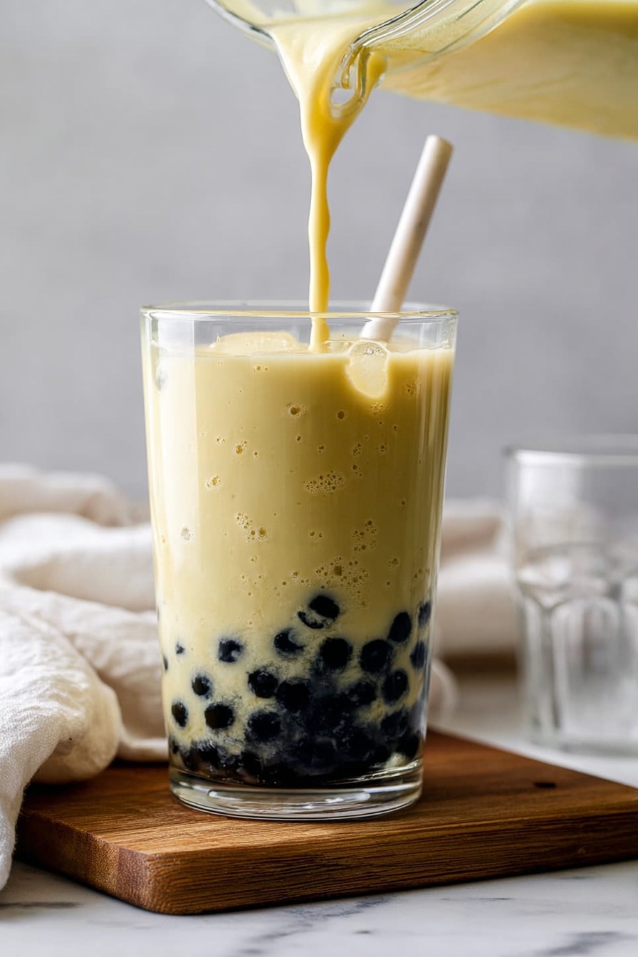 Mango Bubble Tea with Tapioca Pearls, Mango Bubble Tea, Tapioca Pearls Bubble Tea, Fresh Mango Bubble Tea, Homemade Bubble Tea - A clear glass filled with a layer of black tapioca pearls at the bottom, topped with a thick, pale yellow creamy liquid being poured from a blender jar above. The drink has small bubbles on its surface and a smooth texture, and a white straw is inserted into the glass. The glass sits on a small wooden board placed on a white marbled surface, with a blurred white cloth and an empty clear glass in the soft background. Photo taken with an iphone --ar 2:3 --v 7