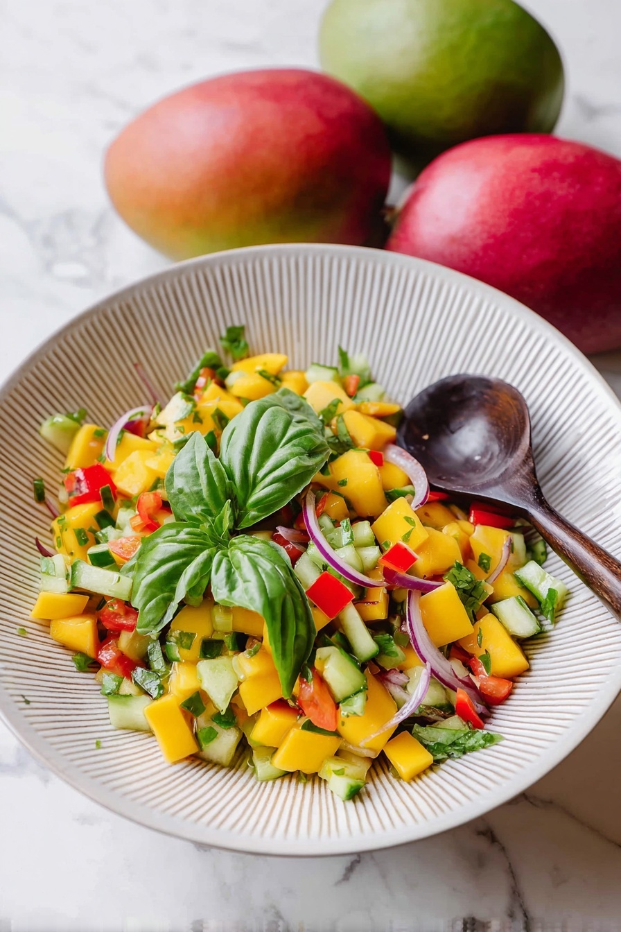 Mango Salad with Lemon-Honey Dressing, mango salad recipe, refreshing fruit salad, easy summer salad, healthy mango salad - A white bowl with fine vertical ridges holds a colorful mango salad. The salad has many small diced yellow mango pieces mixed with green cucumber, red bell pepper chunks, and thin slices of purple onion. There are several fresh green basil leaves on top and mixed throughout the salad. A dark wooden spoon rests inside the bowl on the right side. The bowl is set on a white marbled surface with two whole mangoes, one red and one green, placed slightly blurred in the background. Photo taken with an iphone --ar 2:3 --v 7