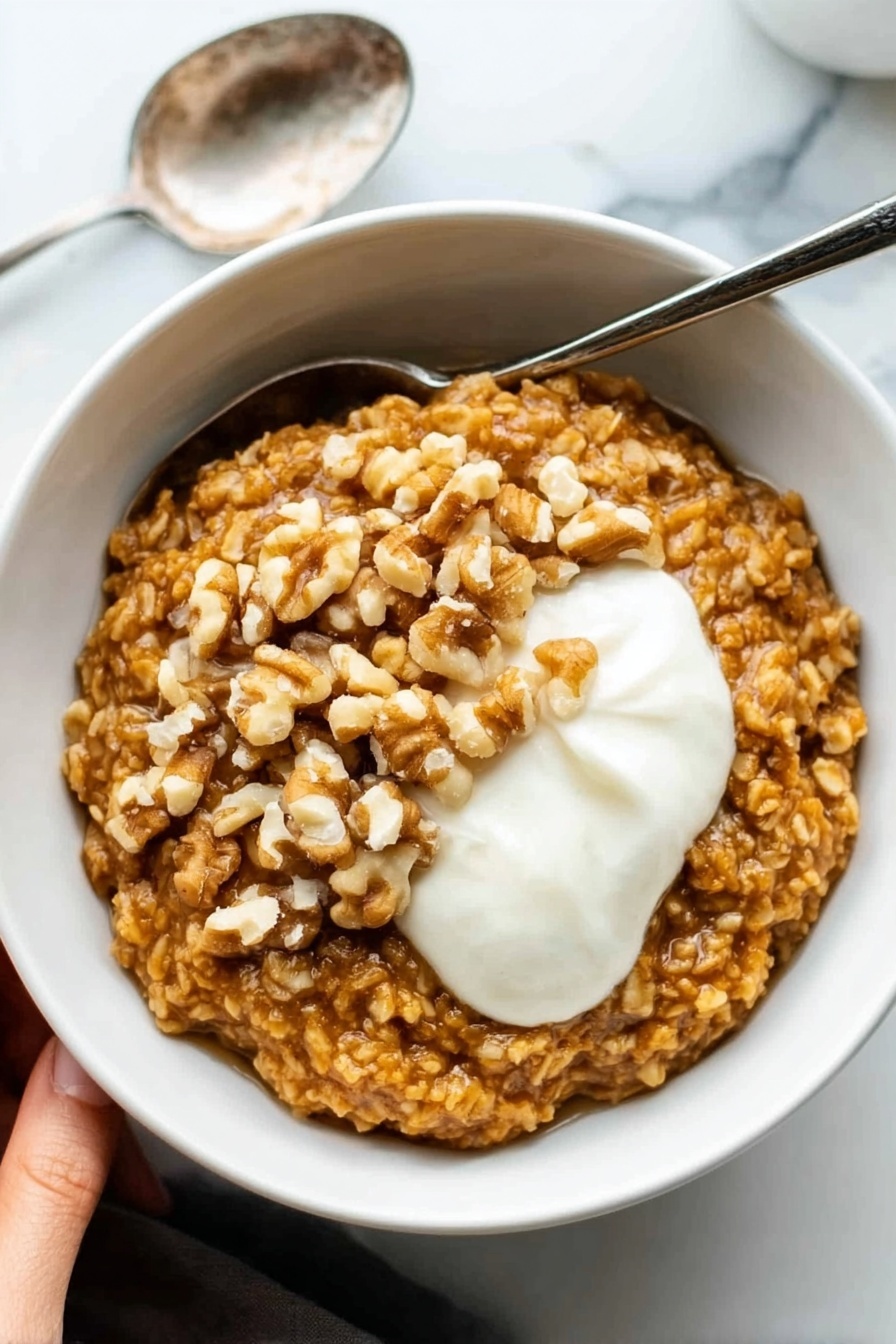 Pumpkin Oatmeal, Pumpkin Oatmeal Recipe, Fall Breakfast Ideas, Cozy Morning Breakfast, Healthy Pumpkin Breakfast - A white bowl filled with thick oatmeal that is golden brown in color and has a textured look from large oats. On top of the oatmeal, there is a layer of whole walnut pieces, adding a rough, uneven surface in light brown shades. Next to the walnuts, there is a dollop of smooth, white cream sitting on the right side of the bowl. The bowl is set on a white marbled surface, and a silver spoon rests next to it on the left. In the upper left corner, a woman's hand is visible holding the bowl. Photo taken with an iphone --ar 2:3 --v 7