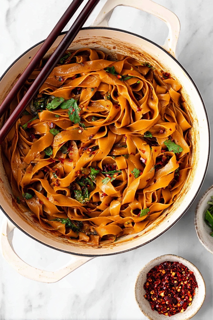 Spicy Chili Garlic Noodles, spicy noodle stir-fry, easy Asian noodle recipe, quick spicy garlic noodles, flavorful chili garlic pasta - A white bowl filled with wide flat noodles coated in a shiny reddish-brown sauce is shown from above. The noodles are mixed and curled inside the bowl with visible pieces of fresh green onions and chopped cilantro scattered on top. There are small white sesame seeds and dark chili flakes sprinkled over the noodles. A pair of dark wooden chopsticks rests on the edge of the bowl leaning inward. In the blurred background, two small white bowls hold red chili flakes and more chopped green onions. The surface is a white marbled texture. photo taken with an iphone --ar 2:3 --v 7
