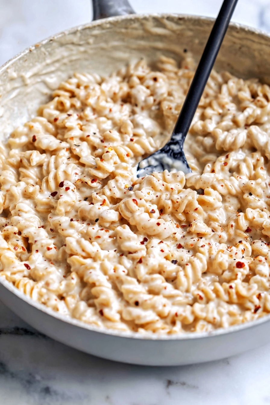 Creamy Cottage Cheese Pasta Sauce, healthy pasta sauce, quick pasta recipe, protein-packed pasta sauce, easy weeknight dinner - The image shows a large gray bowl filled with a creamy, light beige pasta dish. The pasta is rotini, with a spiral shape, fully coated with a thick, smooth sauce that has a cheesy and slightly speckled texture. A woman's hand holding a metal spoon is stirring the pasta, lifting a spoonful from the center. The bowl sits on a surface with a white marbled texture. The overall look is warm and creamy, with the pasta fully covered in the sauce to give it a rich appearance. photo taken with an iphone --ar 2:3 --v 7