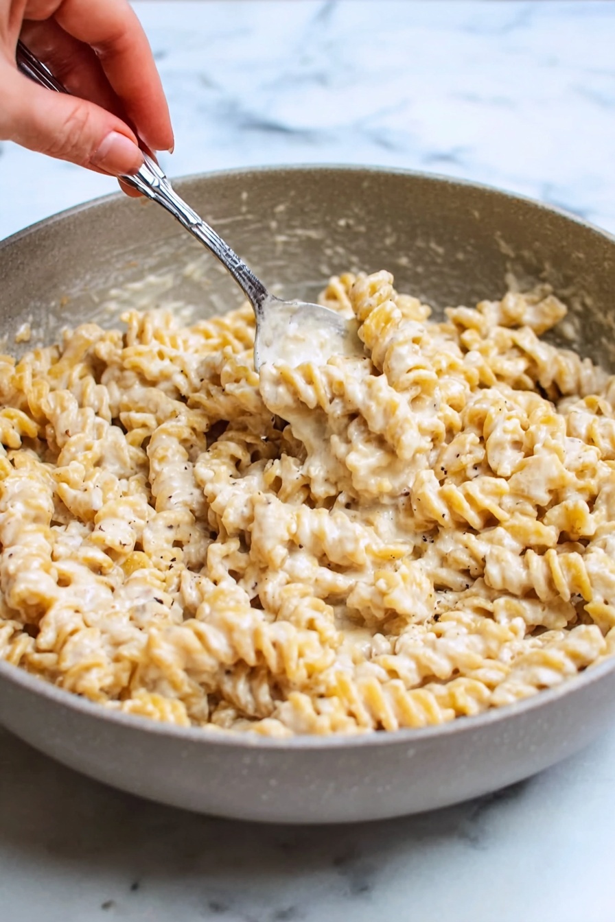 Creamy Cottage Cheese Pasta Sauce, healthy pasta sauce, quick pasta recipe, protein-packed pasta sauce, easy weeknight dinner - A close-up view of a white pan filled with creamy spiral pasta coated in a thick, smooth, off-white sauce with visible small black pepper and red chili flakes mixed in. A black spoon is standing upright in the middle of the dish, slightly submerged in the pasta. The pan sits on a white marbled surface. The overall look is warm and rich with a soft texture on the pasta photo taken with an iphone --ar 2:3 --v 7