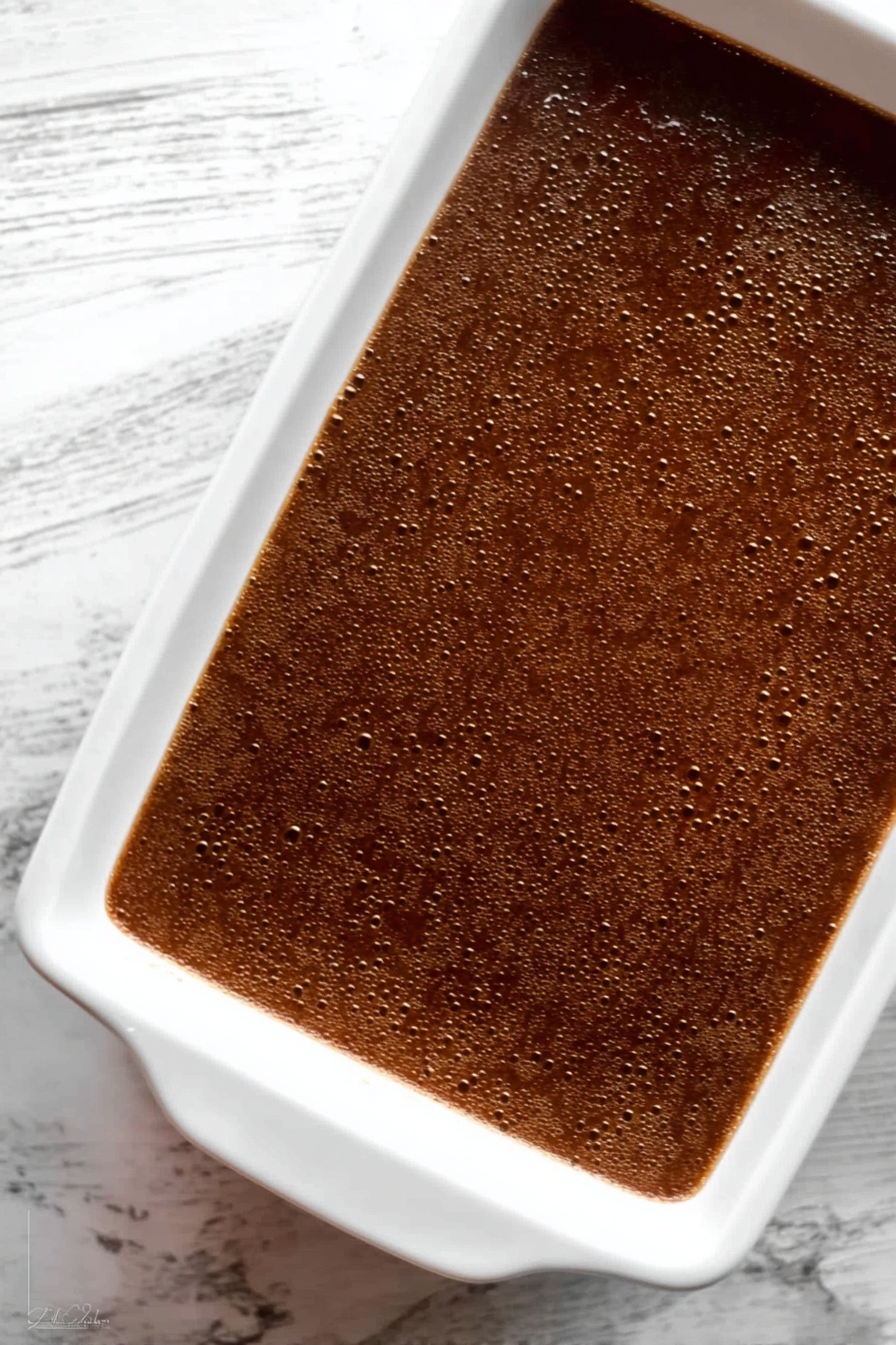 Gingerbread Cake with Butterscotch Sauce, gingerbread cake recipe, spiced gingerbread dessert, moist gingerbread cake, easy holiday gingerbread cake - The image shows a white rectangular baking dish filled with a smooth, dark brown dessert that has a few small bubbles on the surface, giving it a slightly textured look. The dish sits on a white marbled surface with visible veins. The dessert has a shiny, even top layer that reflects some light, indicating a moist texture beneath. Photo taken with an iphone --ar 2:3 --v 7