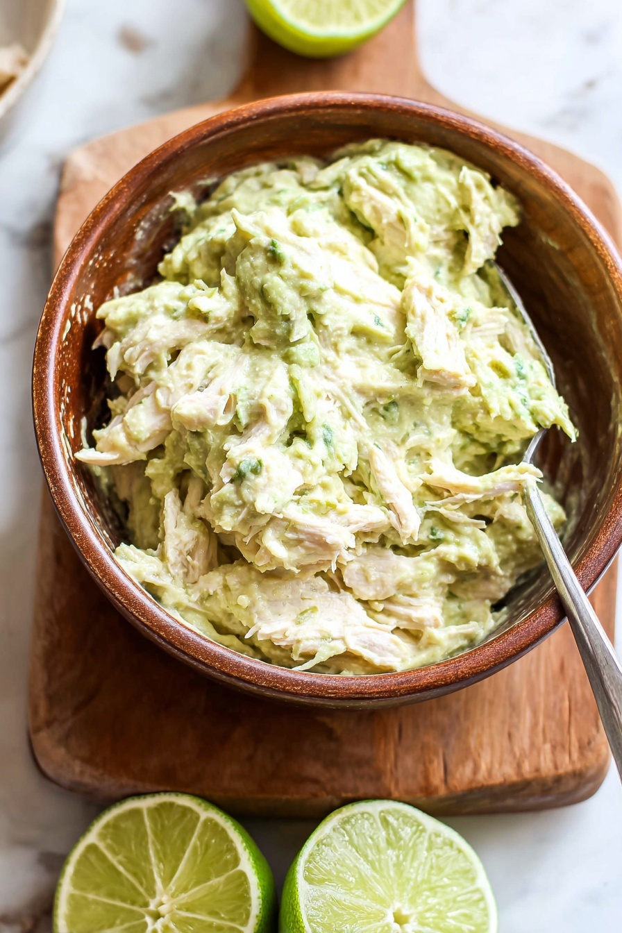 Healthy Greek Yogurt Avocado Chicken Salad, healthy chicken salad, avocado chicken salad, Greek yogurt chicken salad, gluten-free chicken salad - A brown bowl filled with a creamy, light green mixture that has a chunky texture with visible pieces of shredded white meat mixed inside. The bowl is placed on a wooden board, which rests on a white marbled surface. Behind the bowl, there are two halves of a lime, one facing up showing the green juicy interior, and the other turned to the side. A metal spoon is partially inserted into the mixture in the bowl. The scene is bright and fresh. photo taken with an iphone --ar 2:3 --v 7