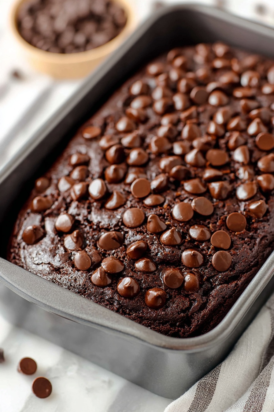 Chocolate Brownie Bread, fudgy chocolate bread, easy chocolate dessert, moist chocolate loaf, chocolate baking ideas - A dark brown chocolate cake with a shiny cracked top fills a grey baking pan. The cake is covered with many round chocolate chips scattered all over the surface, adding texture and a shiny look. The edges of the cake are slightly raised, showing a soft, moist inside. The pan sits on a white marbled surface, with a bowl of chocolate chips blurred out in the background. Photo taken with an iphone --ar 2:3 --v 7