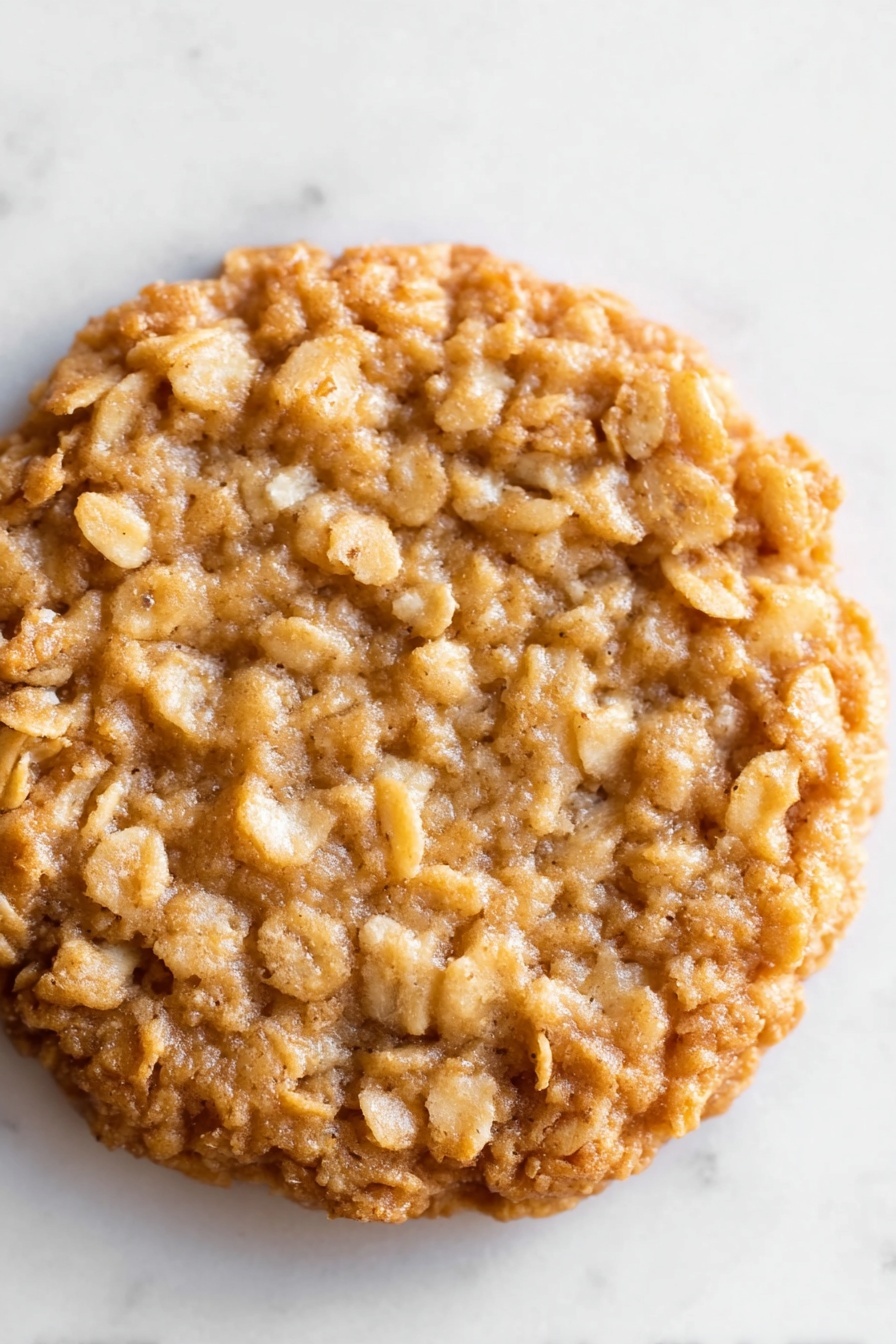 Delicious Lace Cookies with Oats and Brown Butter, lace cookies recipe, oat cookies, brown butter cookies, crispy lace cookies - The image shows a close-up view of a single oatmeal cookie. The cookie is round and thick with an uneven surface made of baked oats. Its color is a warm golden brown with lighter patches of oat flakes throughout, giving it a rough texture. The cookie sits flat on a white marbled surface, and the edges are slightly raised and crispy-looking. There are no other objects or decorations in the frame, focusing all attention on the detailed texture of the oatmeal cookie. photo taken with an iphone --ar 2:3 --v 7