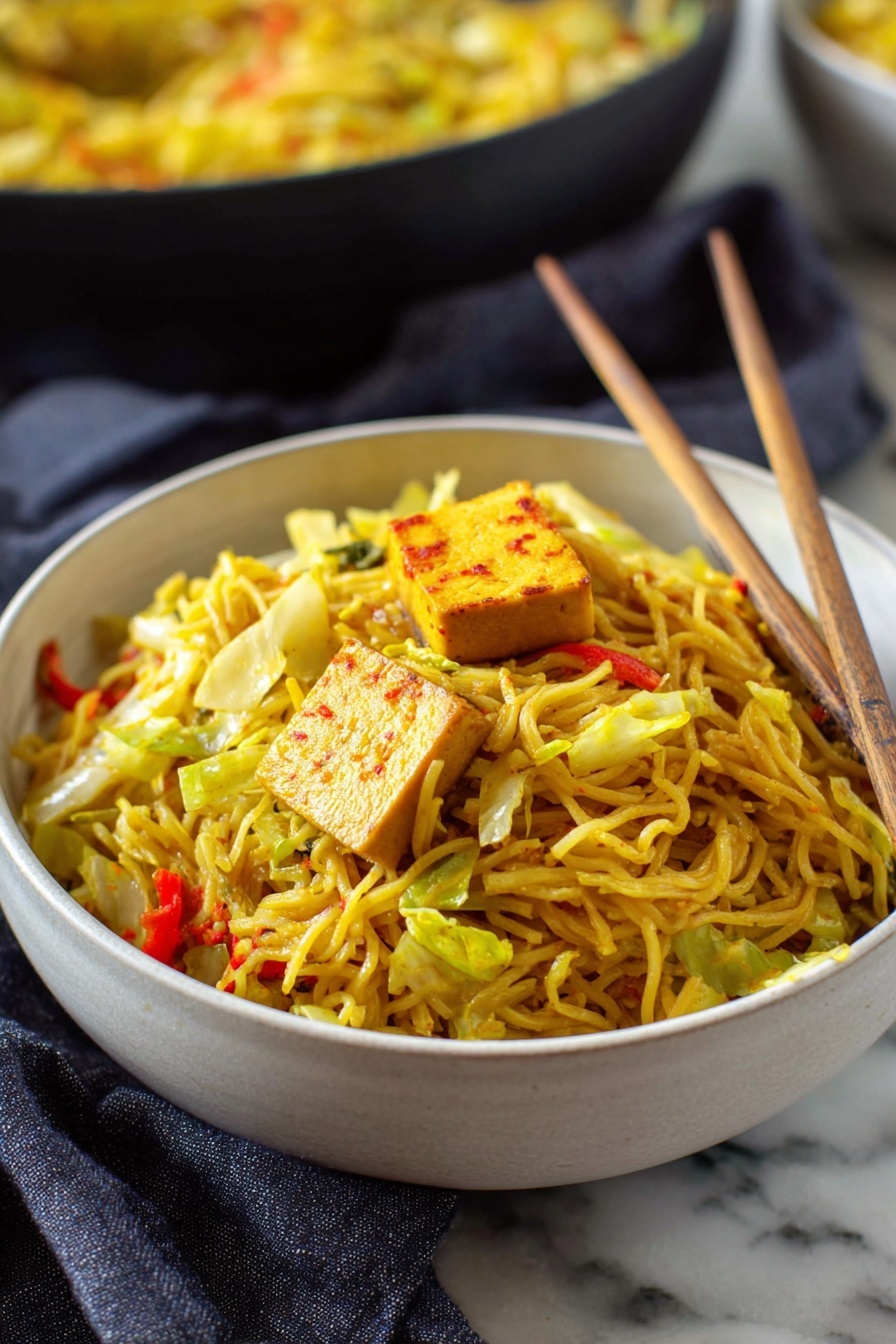 Easy Singapore Noodles, Singapore Noodles recipe, spicy noodle stir-fry, Asian noodle recipe, quick Asian dinners - A deep white bowl full of thin yellow noodles mixed with pieces of green cabbage and small bits of red bell pepper, topped with a square piece of light brown tofu with reddish edges. Light wooden chopsticks rest inside the bowl, leaning against the noodles on the right side. The bowl sits on a white marbled surface with a dark gray cloth nearby. In the background, there is another bowl filled with more noodles, blurred slightly. Photo taken with an iphone --ar 2:3 --v 7