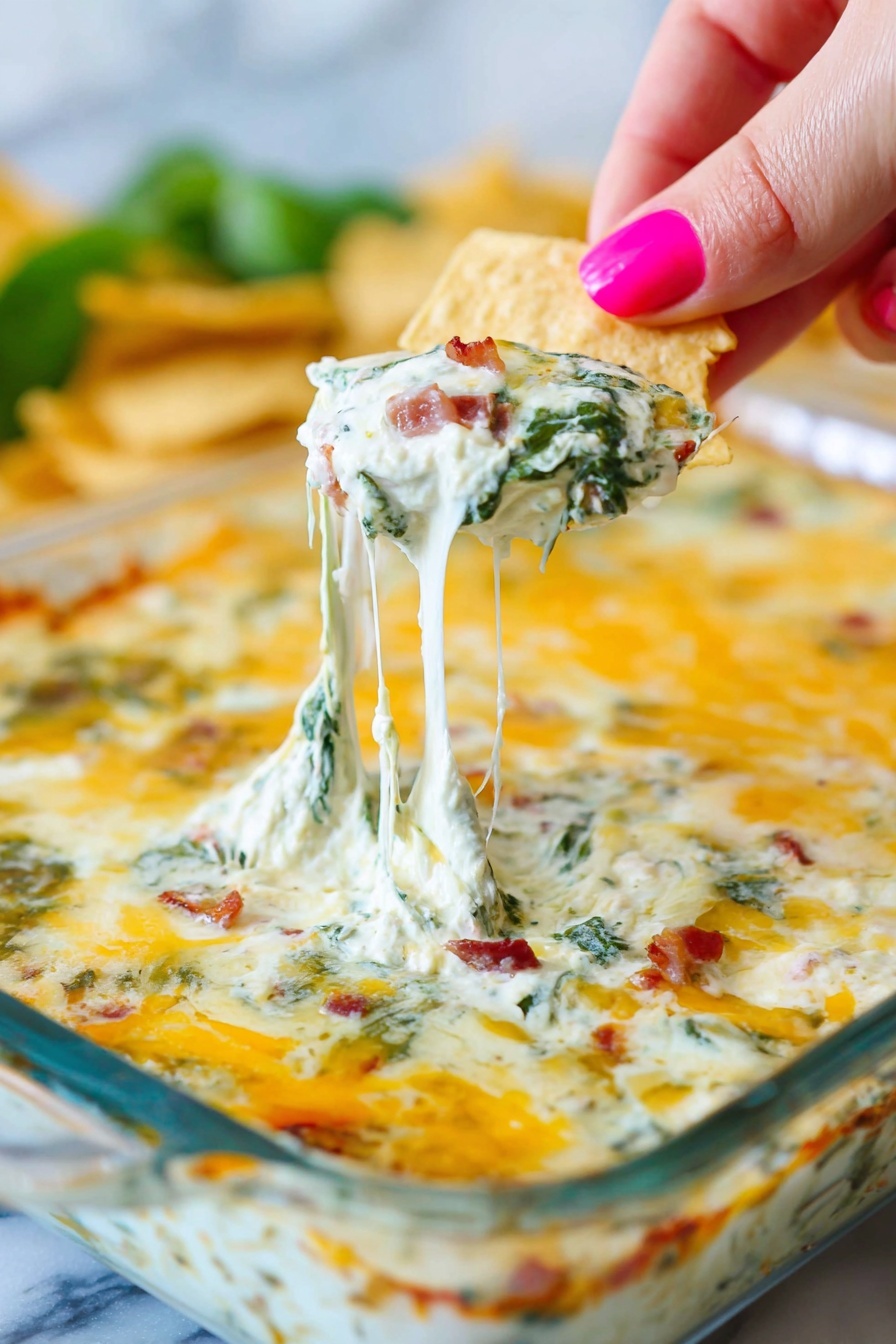 Bacon Spinach Artichoke Dip, cheesy bacon spinach dip, easy bacon appetizer, crowd-pleasing dip recipes, baked spinach artichoke dip - A white square glass dish shows a baked dip with a top layer of melted golden-yellow cheese, lightly browned in spots. Under the cheese, thick creamy white sauce mixed with green spinach leaves and small bits of reddish-brown bacon is visible. A woman's hand with pink painted nails holds a triangular beige chip pulling up a string of stretchy melted cheese from the dish, showing the layers clearly. The background is blurry with hints of green and more chips. The whole scene is set on a white marbled surface. photo taken with an iphone --ar 2:3 --v 7