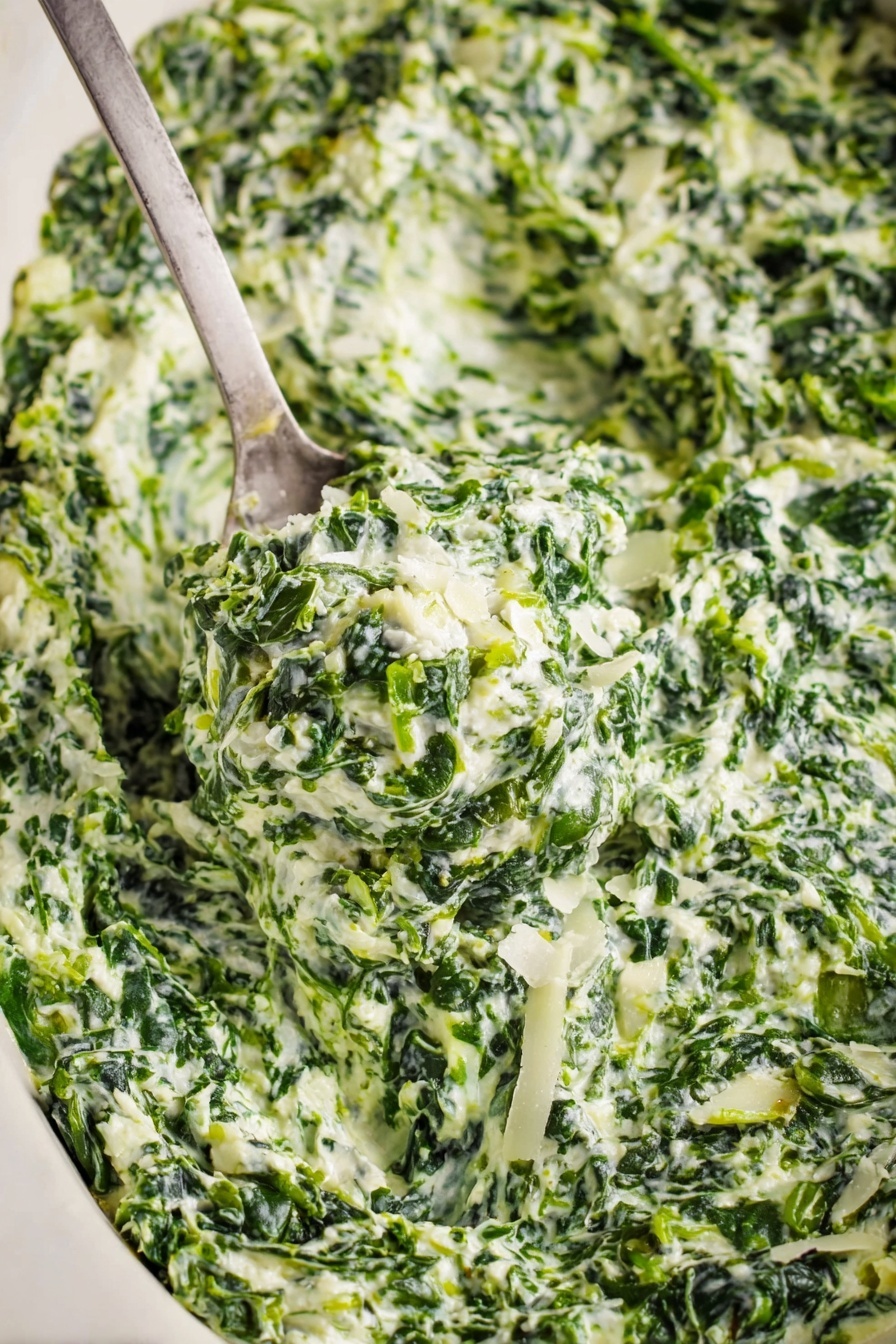 Creamed Spinach, creamed spinach side dish, cheesy creamed spinach, easy creamed spinach recipe, creamy spinach sides - A close-up view of creamy spinach dip with visible layers of finely chopped green spinach mixed with white creamy sauce and small pieces of onion. The texture looks thick and rich with bits of grated white cheese sprinkled on top. A metal spoon scoops up a generous portion of the dip, showing the mix's dense, slightly chunky consistency. The dish sits on a white marbled surface that contrasts with the deep green and white colors of the dip. Photo taken with an iphone --ar 2:3 --v 7