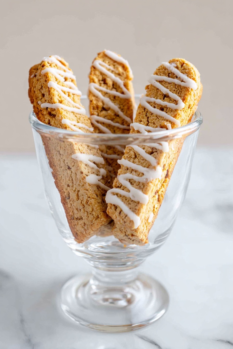Cinnamon Vanilla Biscotti, best biscotti recipe, crunchy biscotti with cinnamon, homemade vanilla biscotti, coffee pairing biscotti - The image shows a clear glass cup placed on a white marbled surface. Inside the cup, there are three crispy biscotti standing upright, each with a rough light brown texture. White icing is drizzled in thin stripes across each biscotti, adding contrast to their warm golden color. The background is plain and softly lit, emphasizing the cup and its contents. photo taken with an iphone --ar 2:3 --v 7