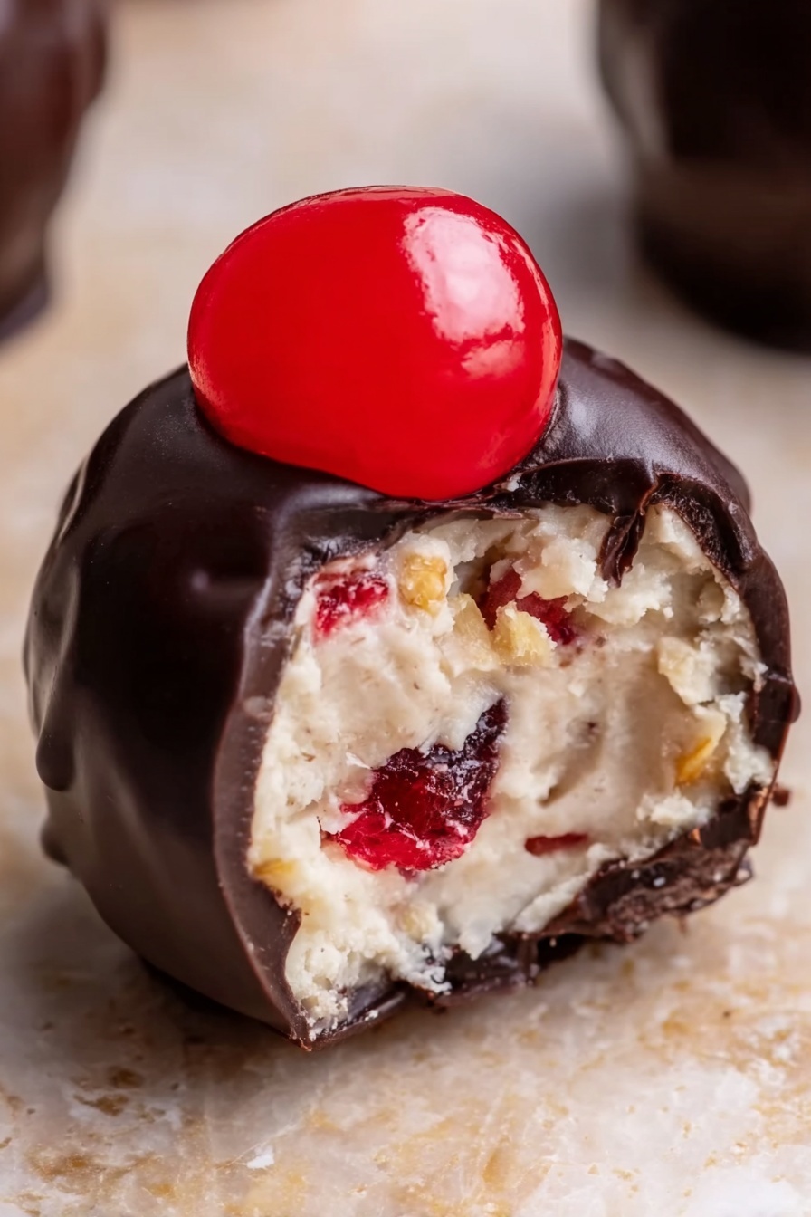 Cherry Bourbon Balls, bourbon cherry treats, chocolate cherry candies, adult holiday desserts, easy bourbon ball recipes - The image shows a close-up view of a small dessert ball with three layers. The outside layer is dark, smooth, and shiny chocolate coating covering the entire ball. Inside, the second layer is light-colored, creamy, and soft with some small red and golden brown bits mixed inside, giving it a textured look. On top of the chocolate layer, there is a bright red, glossy half of a cherry placed in the center. The ball sits on a light brown surface with a white marbled texture softly blurred in the background. photo taken with an iphone --ar 2:3 --v 7