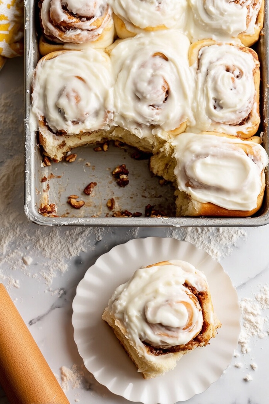Soft Apple Cinnamon Rolls, Apple Cinnamon Rolls, Homemade Cinnamon Rolls, Fluffy Cinnamon Roll Recipe, Autumn Breakfast Treats - A metal baking dish holds six soft cinnamon rolls, with one removed showing the bottom layer coated in a sticky cinnamon filling and bits of nuts. Each roll has a creamy white icing thickly spread on top, swirled smoothly over the soft golden brown dough, with visible cinnamon spirals peeking through under the icing. One cinnamon roll sits on a small white scalloped plate in the lower right corner, showing its fluffy layered inside. The background is a white marbled surface lightly dusted with flour, with a wooden rolling pin nearby. photo taken with an iphone --ar 2:3 --v 7