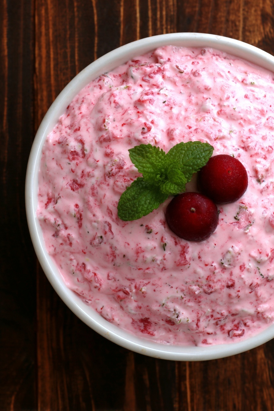 Cranberry Fluff Salad, holiday side recipes, festive Christmas salads, easy cranberry salad, creamy fruit salad - The image shows a white bowl filled with a thick, creamy pink mixture that has small bits of darker pink throughout. On top of the mixture, there are two small red berries and a small green mint leaf. The bowl is sitting on a dark wooden surface. photo taken with an iphone --ar 2:3 --v 7