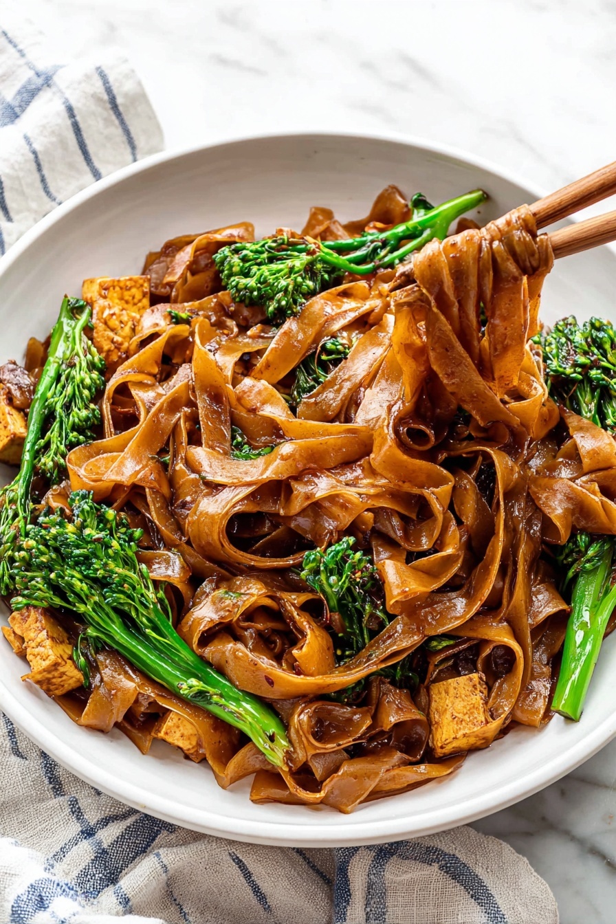 Quick Thai Pad See Ew Stir Fry, Thai noodle stir fry, easy Pad See Ew recipe, Thai street food at home, quick weeknight Thai dinner - A white bowl is filled with wide, flat noodles coated in a shiny dark brown sauce, layered thick, with some noodles lifted by wooden chopsticks to the right side of the image. Mixed in are pieces of light brown cooked tofu and bright green broccolini with textured small florets. The bowl sits on a white cloth with blue stripes on a white marbled surface. The colors show a contrast between the dark noodles, green vegetables, and pale tofu, creating a rich and fresh look. photo taken with an iphone --ar 2:3 --v 7