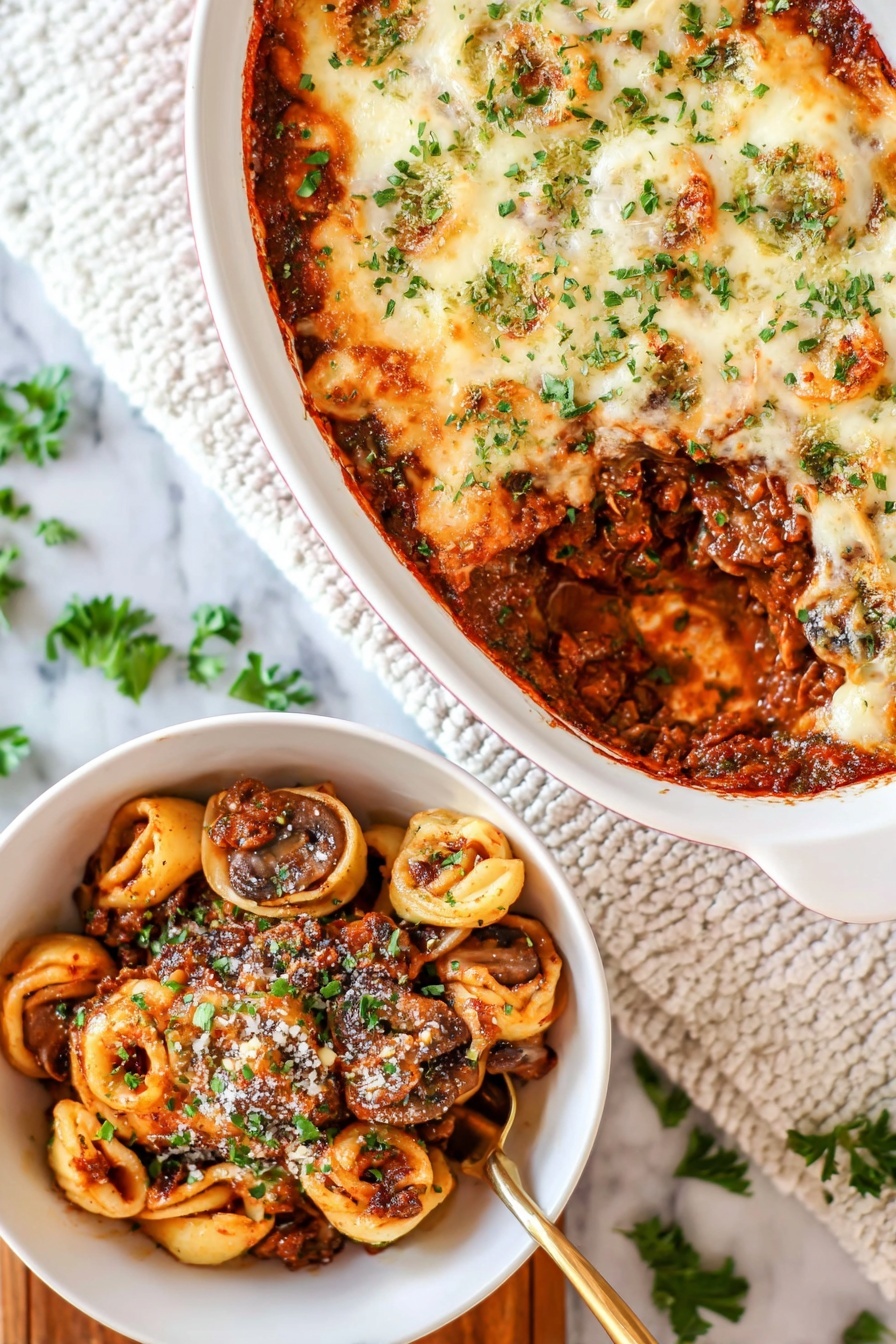 Cheesy Baked Tortellini with Beef, baked tortellini with beef, cheesy stuffed pasta bake, easy cheesy beef casserole, comfort food dinner recipes - The image shows a white oval baking dish with a layered casserole on a white marbled surface. The top layer is melted, lightly browned cheese sprinkled with green herbs. A section is scooped out, revealing a rich, dark brown meat sauce mixed with whole mushrooms and tortillas shaped like small circles inside the dish. Next to it is a white bowl filled with a serving of the casserole, showing layers of browned meat sauce, mushrooms, and pasta circles, all topped with some grated cheese and chopped green herbs. A golden spoon is partially visible in the bowl. The background includes a white textured cloth and scattered green herb leaves. photo taken with an iphone --ar 2:3 --v 7