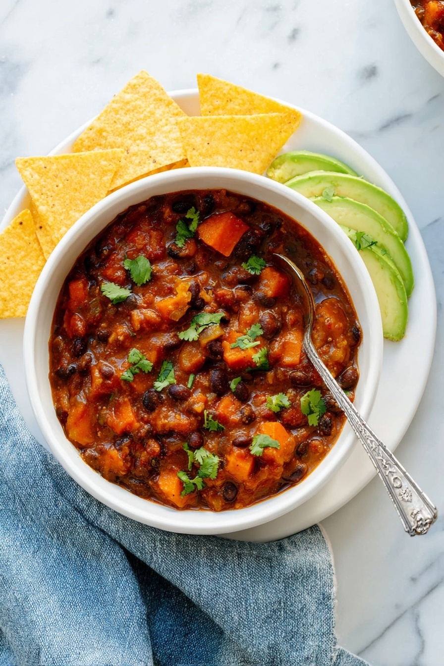 Vegetarian Chili, vegetarian chili recipe, plant-based chili, healthy vegetarian dinner, quick vegetarian chili - A white bowl filled with thick chili containing black beans, orange carrot pieces, and chunks of sweet potato in a reddish-brown sauce, garnished with green cilantro leaves. On the right side inside the bowl are thin slices of light green avocado sprinkled with cilantro. The bowl sits on a white plate arranged with five triangular yellow tortilla chips around the left side. A silver spoon with a decorative handle rests inside the chili. The setting has a white marbled texture surface with a slightly crumpled blue cloth napkin next to the bowl. Photo taken with an iphone --ar 2:3 --v 7