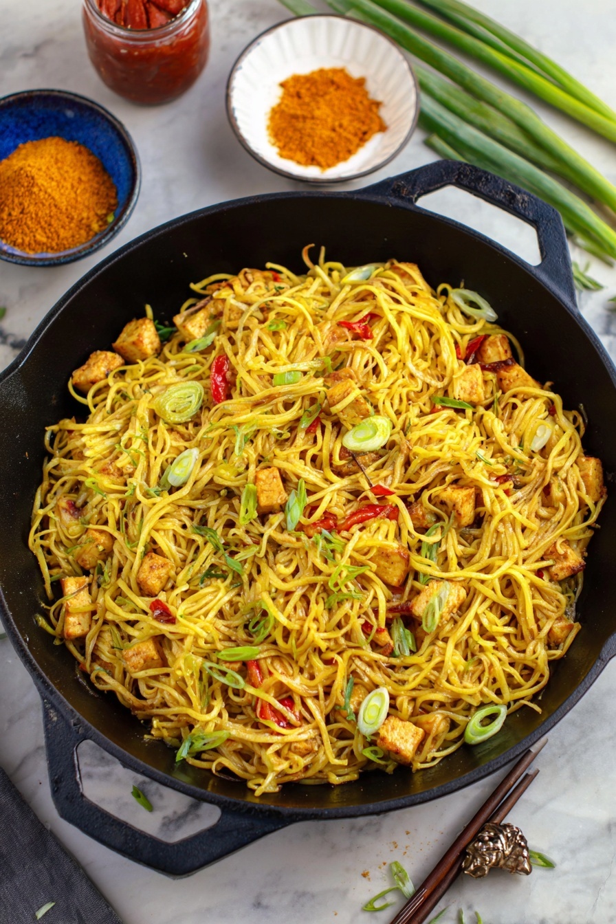 Easy Singapore Noodles, Singapore Noodles recipe, spicy noodle stir-fry, Asian noodle recipe, quick Asian dinners - A black cast iron pan filled with several layers of yellow noodles mixed with small pieces of grilled tofu that show a light brown char, scattered with thin slices of green onions and small bits of red bell pepper. The noodles have a slight shine and look seasoned. Around the pan, there is a white bowl with orange powder, a jar with red sauce, a blue bowl with chopsticks placed on the side, and some green onion stalks on a white marbled surface. The scene has a mix of greens and yellows that make the dish look fresh and tasty photo taken with an iphone --ar 2:3 --v 7
